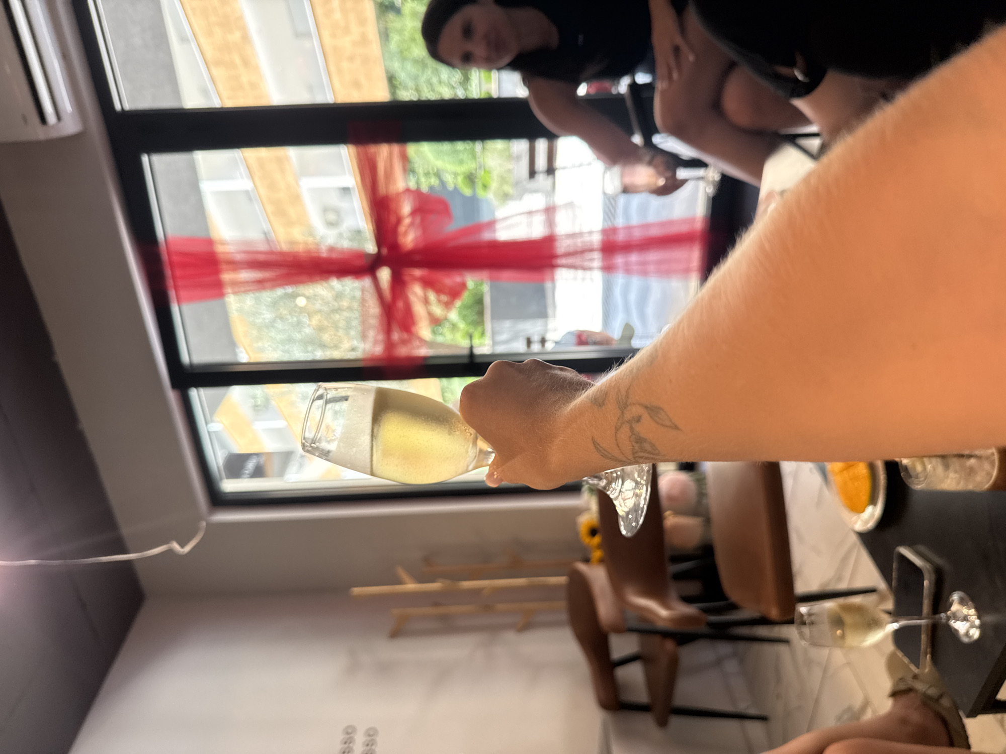 The image depicts a group of people in what appears to be a café or restaurant in Budva, Montenegro, possibly celebrating an event given the prominent red ribbon decoration on the window. In the foreground, a hand is holding up a glass of champagne, suggesting a toast or celebration. The background shows other individuals seated, with one woman in a black dress clearly visible. The setting includes indoor furniture like chairs and tables, as well as an outdoor view through a large window, hinting at the urban environment of Budva. The overall atmosphere seems festive and social.