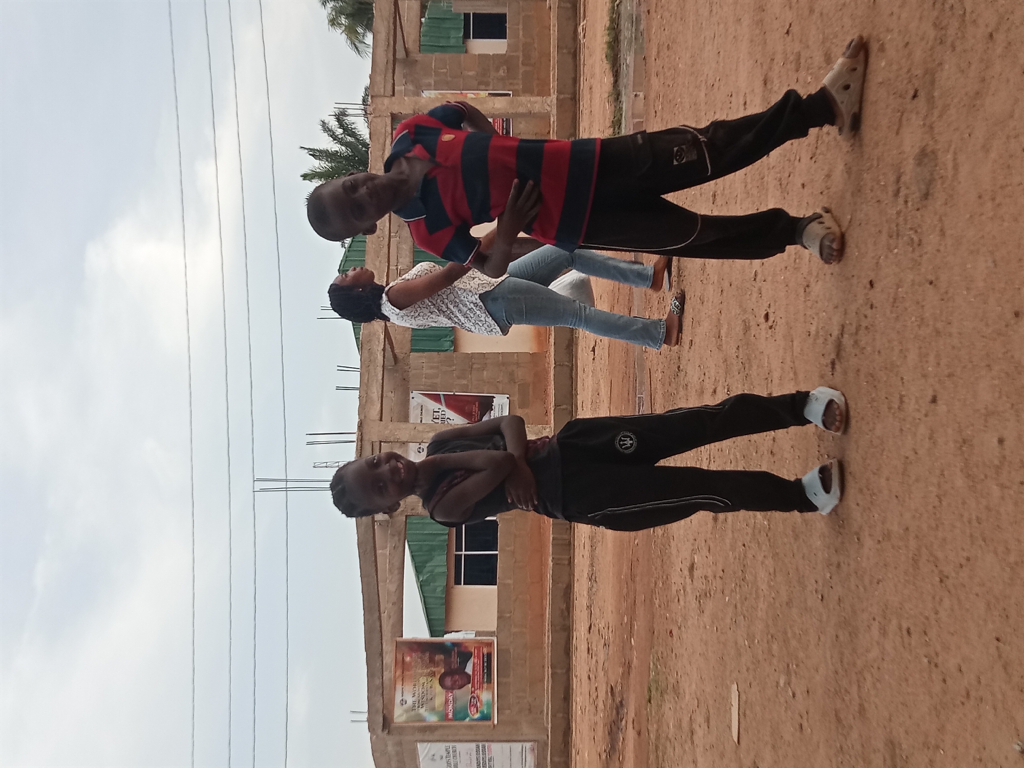 Three people are outside a building in Sabon Tasha, Nigeria. A young woman stands with her arms crossed and smiles at the camera. Next to her, a couple, a man and woman, are standing together. The man has his arms around the woman. The building behind them has posters on it. Overhead are power lines. The ground is reddish-brown dirt.