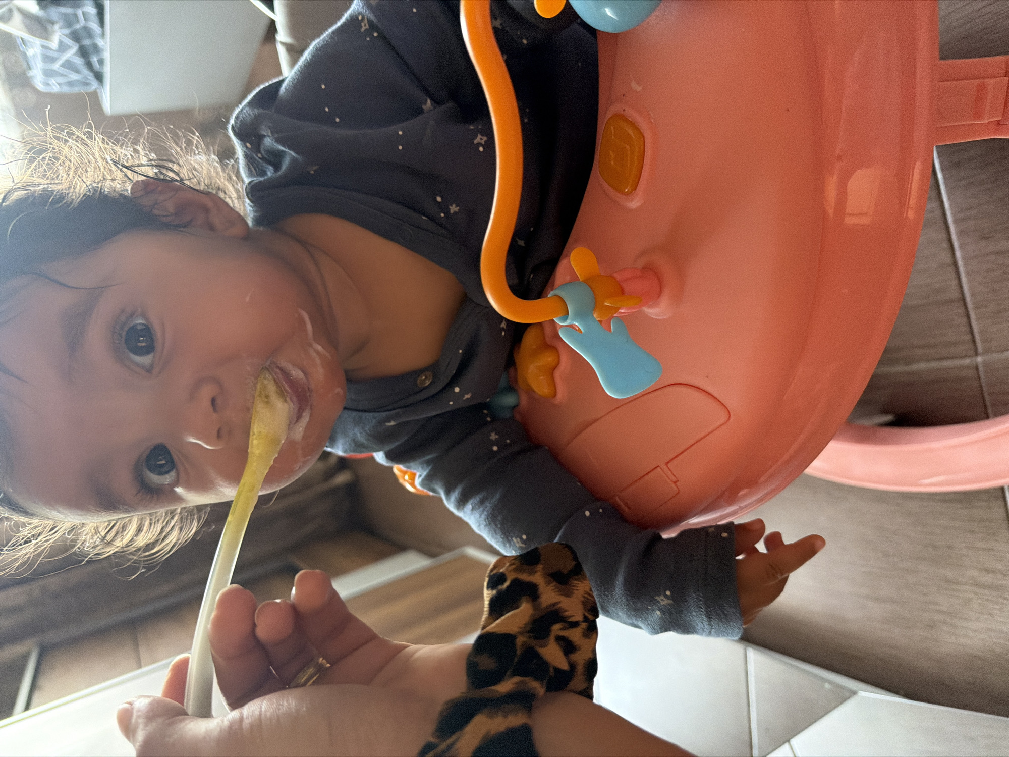 A close-up, eye-level shot shows a baby girl being fed with a spoon. She is wearing a dark blue shirt with white polka dots and is seated in a pink baby walker with orange and blue toys. The baby has dark hair and large, expressive eyes. A hand, possibly belonging to a caregiver, is visible on the left, holding a yellow spoon with food, which is being offered to the baby. The background is blurred, but appears to be an indoor setting with wooden floors and a hint of furniture. The lighting suggests a bright environment, with light catching the baby's hair and creating a halo effect. The overall scene conveys a moment of feeding and care. The location is Razhevo Konare, Bulgaria.