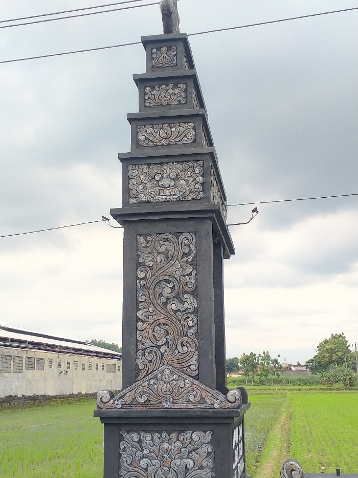 The image displays a tall, dark grey decorative gate pillar or architectural column, likely made of concrete or stone. The pillar is intricately carved with traditional Indonesian motifs. A prominent feature is a stylized, somewhat fierce face with large eyes and a wide mouth, possibly representing a Boma or Kala face, located on one of the upper tiers. Below and around this, elaborate swirling floral and foliate patterns are carved into the pillar, rendered in lighter grey and brownish tones against the darker background of the pillar's surface.
In the background to the left, a long, low-lying building with a light-colored roof and numerous small rectangular windows or vents is visible. Lush green rice paddies or agricultural fields dominate the foreground and mid-ground on both sides of the pillar, with a narrow dirt path running through the fields on the right. Further in the distance on the right, trees and some indistinct buildings are present under an overcast, cloudy sky. Electrical wires stretch horizontally across the upper portion of the frame, passing near the top of the pillar. A small, dark bird can be seen perched on a branch or wire near the pillar in some cropped views.
The scene depicts a static, rural landscape in Indonesia (Kecamatan Kebakkramat), characterized by traditional architecture and agriculture. The time of day appears to be daytime, with soft, diffused light due to the cloudy weather. No people or activities are visible, and there is no discernible text in the image.