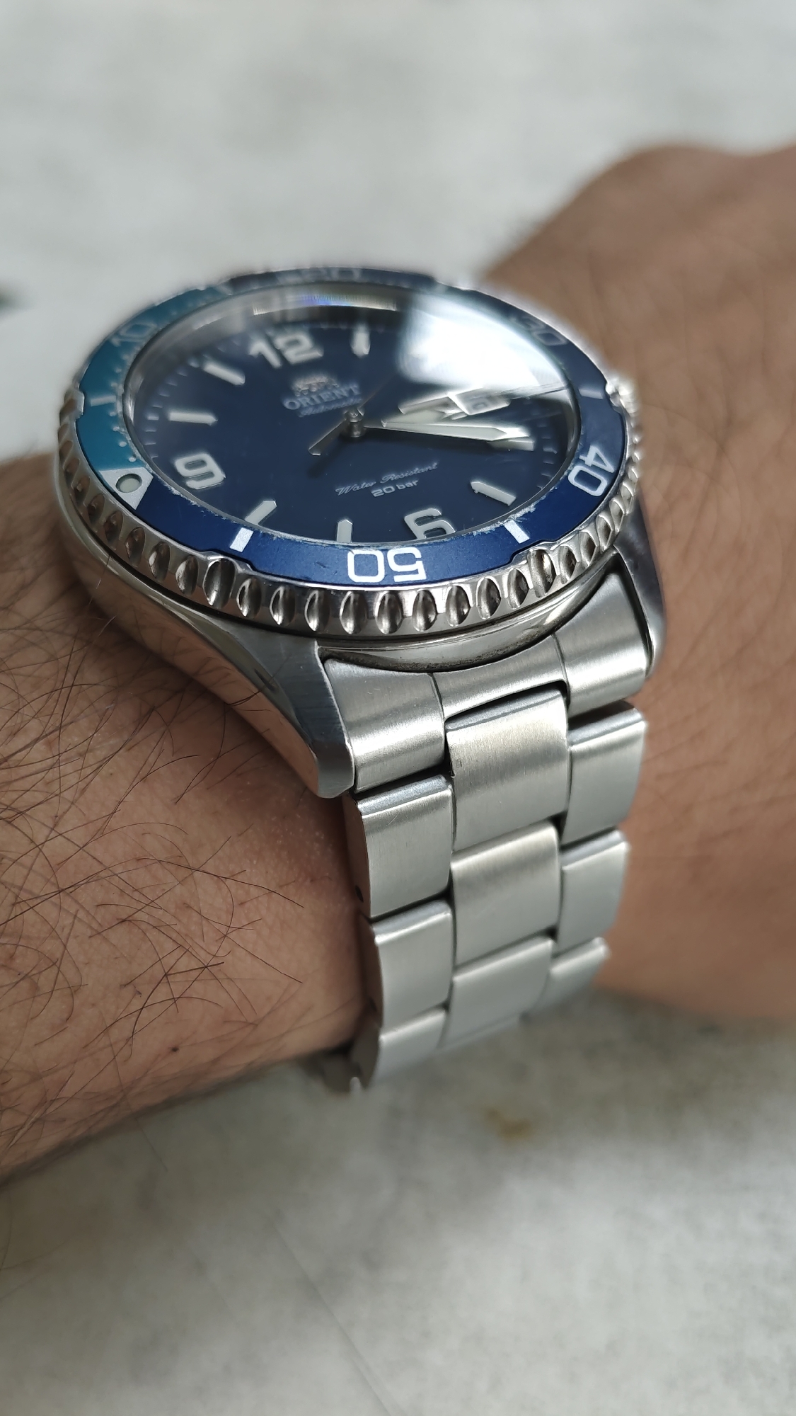 A close-up, high-angle shot shows a stainless steel Orient brand dive watch worn on a person's wrist. The watch features a blue dial with silver hour markers and hands, a unidirectional blue and light blue bezel with white minute markers, and a stainless steel link bracelet. The watch is positioned on the wrist with the dial facing upward, and the bracelet is secured. The background is out of focus, showing a light-colored surface, possibly a table. The image does not depict the city of Karabash, Russia.