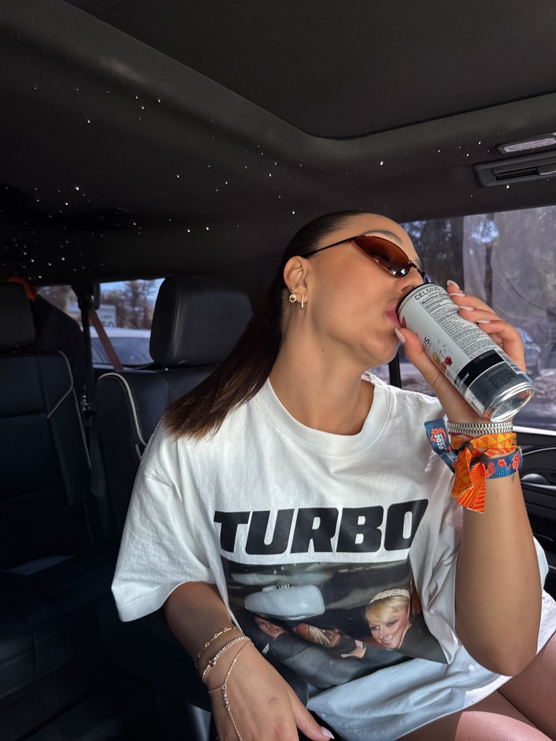 A woman with dark hair styled in a sleek ponytail is depicted from the chest up, seated inside a car, likely in the passenger seat. She is actively drinking from a silver and white beverage can, holding it to her lips. She wears small, dark brown cat-eye sunglasses and a collection of delicate, gold-toned bracelets on her left wrist and hand, one of which extends to her finger. Small gold earrings are visible on her left ear.
She is dressed in a white, oversized t-shirt that features "TURBO" in large black capital letters across the chest. Below this text is a black and white image showing two people inside what appears to be a car, with a blonde woman wearing a tiara prominently smiling towards the viewer. On her right wrist, several colorful fabric wristbands are worn, suggesting attendance at an event, with one blue and orange band displaying the year "2023."
The can she is drinking from clearly shows the brand name "CELSIUS" in black text, along with "Nutrition Facts" and "calories" printed on its side. The interior of the car is dark, featuring visible headrests and a distinctive dark headliner embedded with numerous small, illuminated points, creating a "starry night" effect. Bright, natural daylight streams into the vehicle, indicating it is daytime. The scene conveys a casual and relaxed atmosphere.