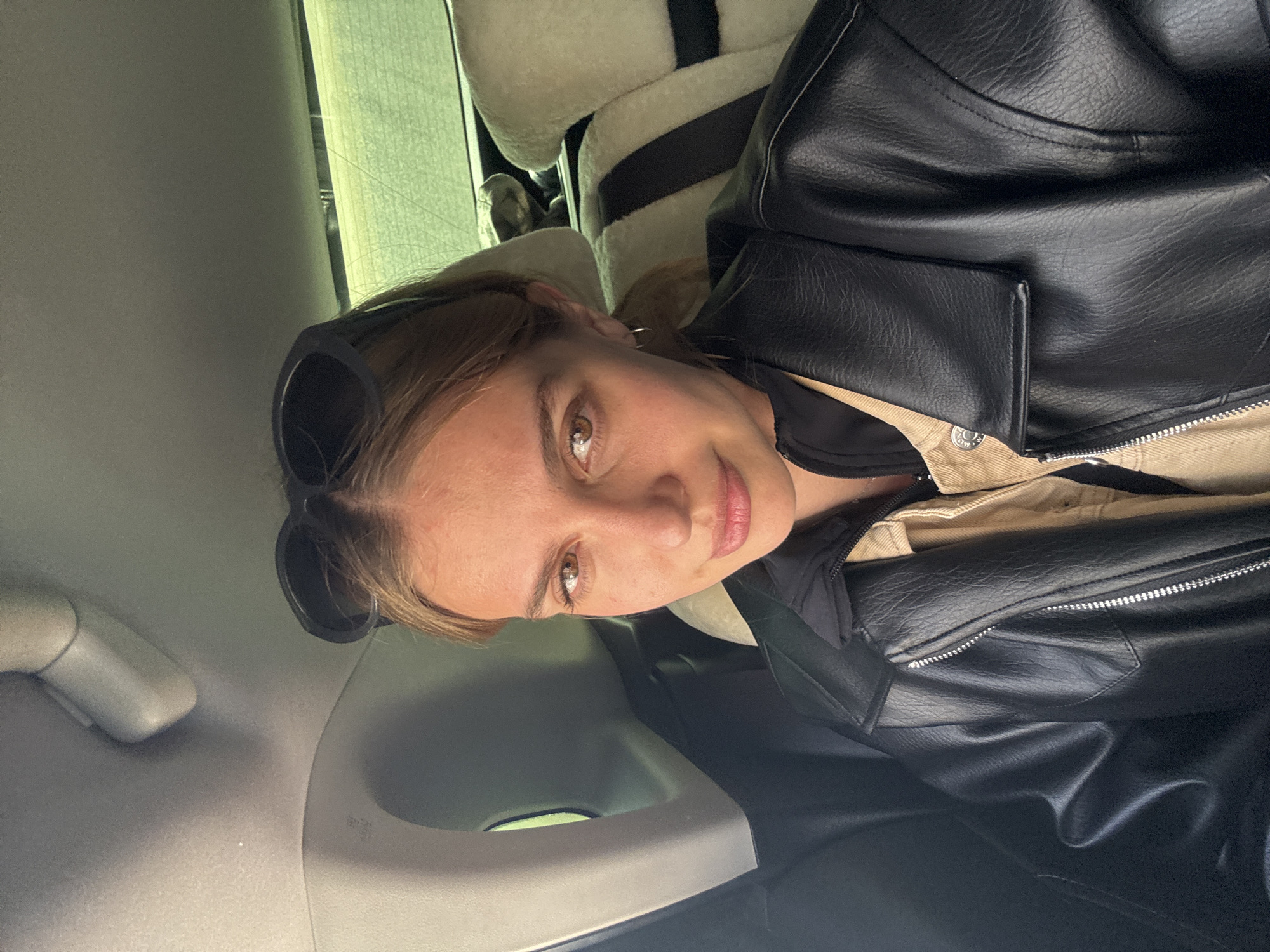 A fair-skinned woman with light brown hair is seated in a car. She is wearing a black leather jacket over a tan collared shirt. Sunglasses are perched on her head. The interior of the car features light gray upholstery and sheepskin seat covers. The location is not discernible to be within Yekaterinburg, Russia.