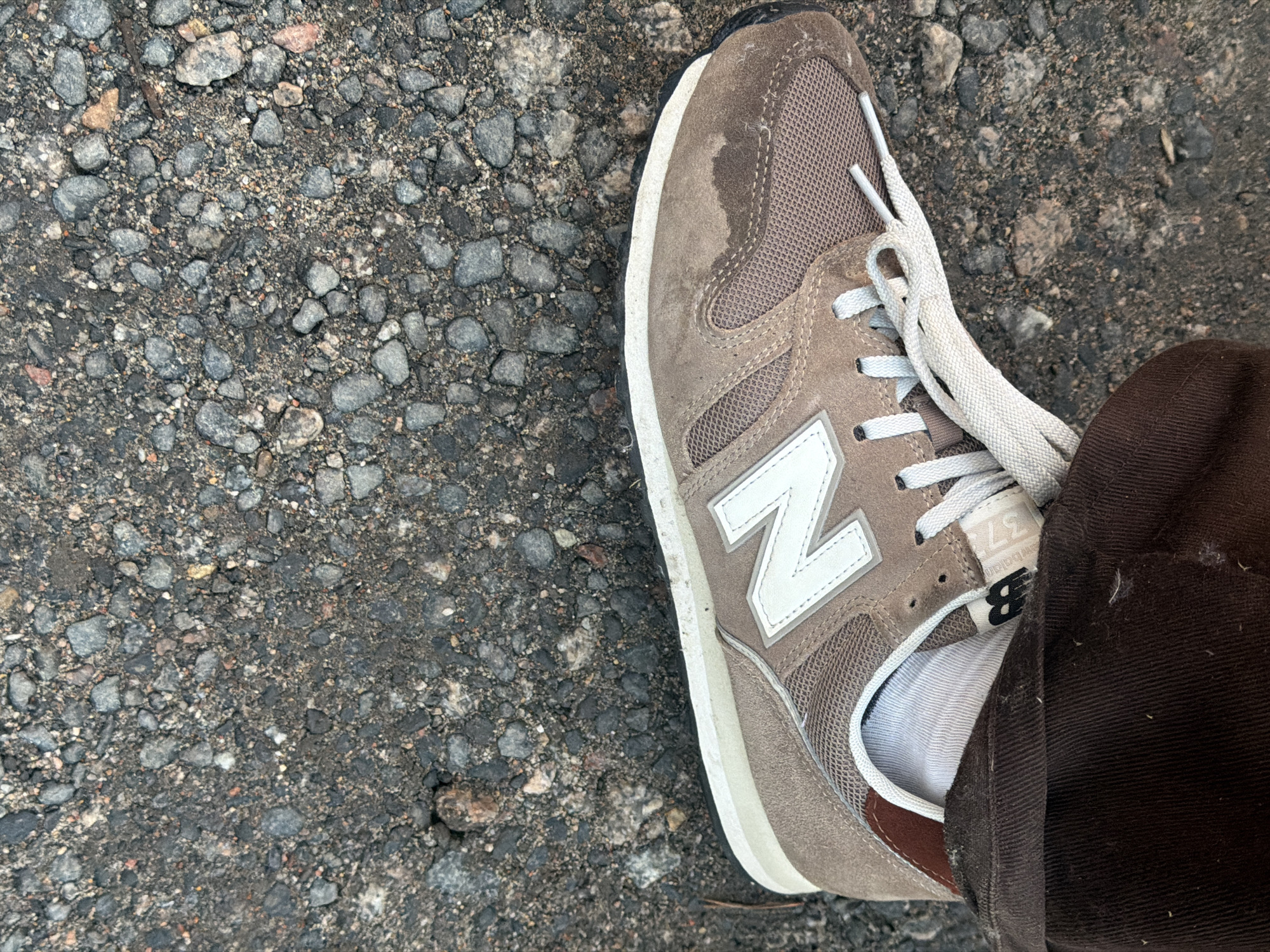 The image shows a close-up, overhead view of a person's foot wearing a brown New Balance sneaker. The sneaker has a white "N" logo on the side and white laces. The foot is clad in a white sock and brown pants. The sneaker rests on a surface of dark asphalt mixed with small gray and reddish-brown stones. The background is entirely composed of this rocky asphalt. There is a wet patch on the upper portion of the sneaker.