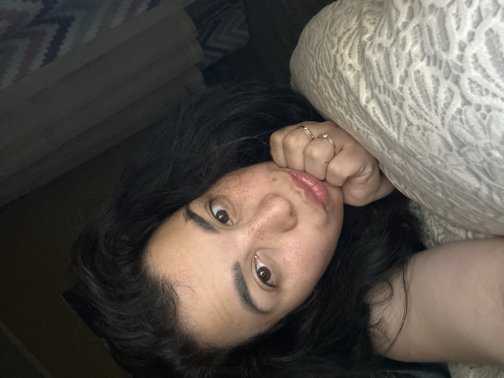 A woman with dark, wavy hair and brown eyes is lying down in bed, resting her head on her hand. She is wearing two rings on her right hand, one of which has a small diamond. Her lips are slightly parted, and she appears to be looking at the camera. The background is dark, with a patterned blanket visible on the right side of the image. The lighting suggests it is either early morning or late at night.