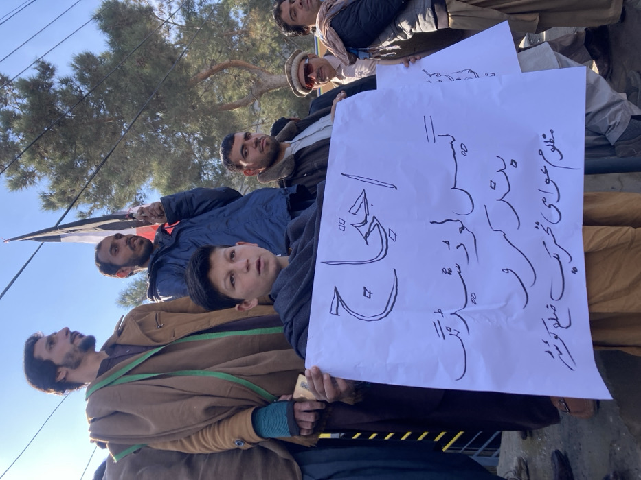 The image depicts an outdoor gathering of approximately six to seven males, including one juvenile, participating in a public assembly or protest in Quetta City Tehsil, Pakistan.
In the foreground, a juvenile male, estimated to be in his early teens, with short dark hair, is positioned centrally, holding a white protest sign. He wears a dark grey shawl draped over a light brown traditional garment. The sign displays black Urdu script, which translates to: "Protest," "Stop Gas Loadshedding," and "Oppressed Public Movement Quetta District."
To the left and slightly behind the juvenile, an adult male with a beard wears a brown jacket over a dark shirt, with a green fabric piece draped over his left shoulder. He holds a light-colored object, possibly a mobile phone, in his left hand. Directly behind the juvenile, another adult male with a beard, wearing a dark blue jacket, holds a flag featuring horizontal stripes of black, white, and red. To the immediate right of the juvenile, an adult male with a beard is visible, wearing a dark brown jacket with a prominent fur collar over a light-colored shirt. Further right, another adult male wears sunglasses and a light-colored traditional cap (Pakol).
The environment includes a clear blue sky overhead with visible utility wires. In the background, green trees are present, suggesting a park-like or roadside setting. The visible ground surface is light-colored concrete or pavement. The individuals are standing in a grouped formation, indicative of an organized public demonstration.