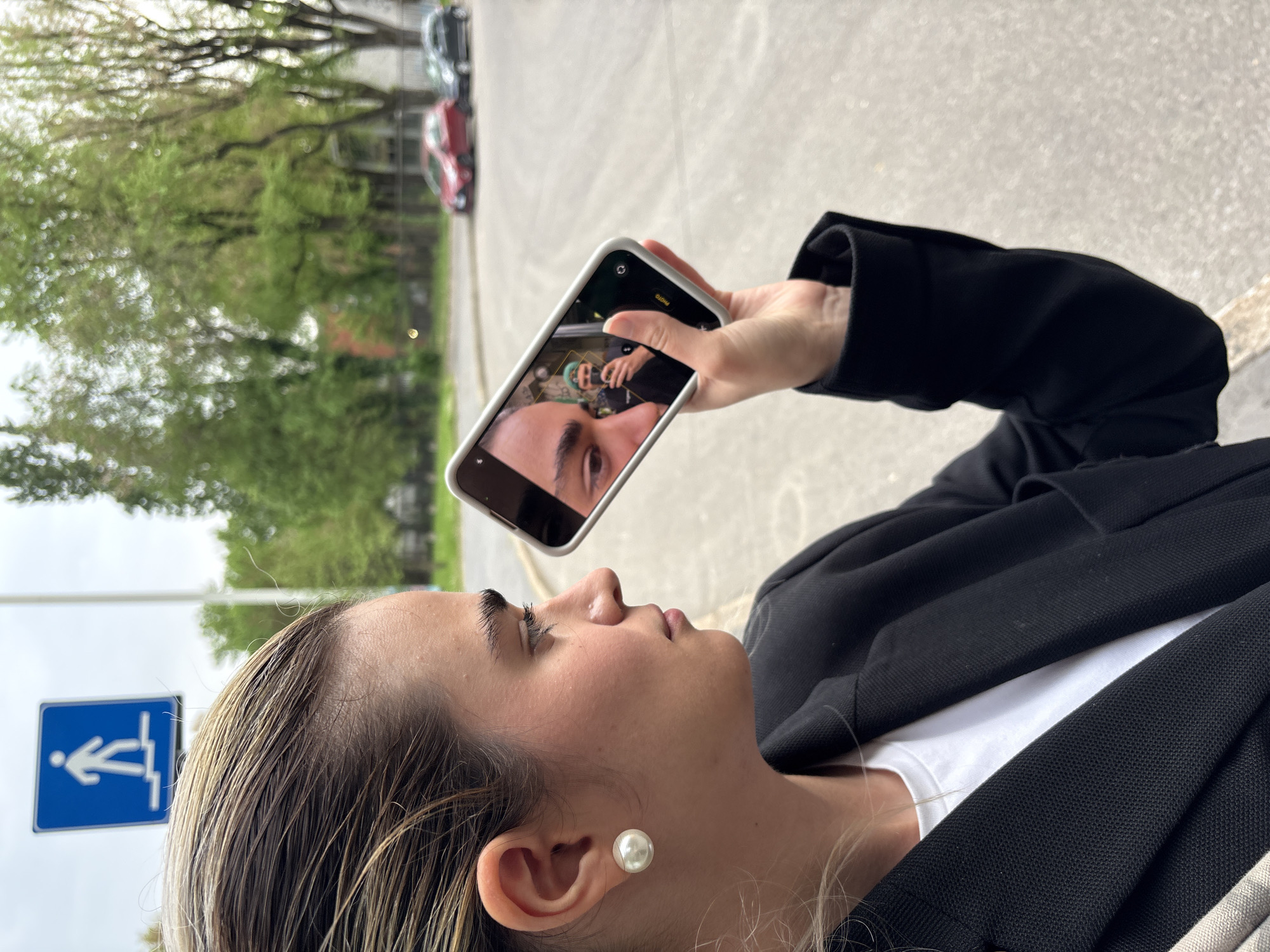 A woman is holding a smartphone, displaying a selfie of another person, in front of her face outdoors. The woman wears a black blazer, white top, and a pearl earring. Her hair is light brown and wet, slicked back. A blue handicapped access sign is visible to the left. The background includes blurred cars and green trees. The scene appears to be on a city street, although specific details confirming the location as Milan, Italy are not directly discernible from the visual information provided.