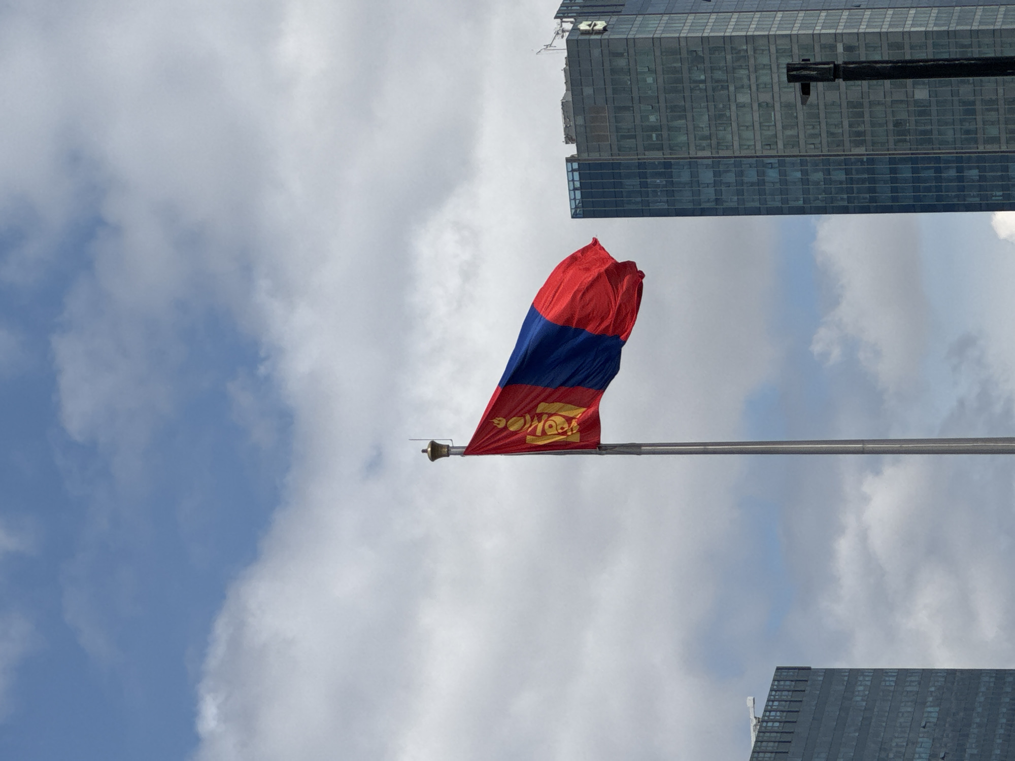 The flag of Mongolia waves prominently in the foreground, its red and blue stripes distinct against a backdrop of a partly cloudy sky. The flag's Soyombo symbol is visible on the red stripe nearest the pole. The flag is mounted on a horizontal pole, seemingly metal, that stretches across the middle of the frame. In the background, sections of modern skyscrapers rise on either side of the flag, their glass facades reflecting the sky. The buildings indicate an urban environment, specifically in Ulaanbaatar, Mongolia.