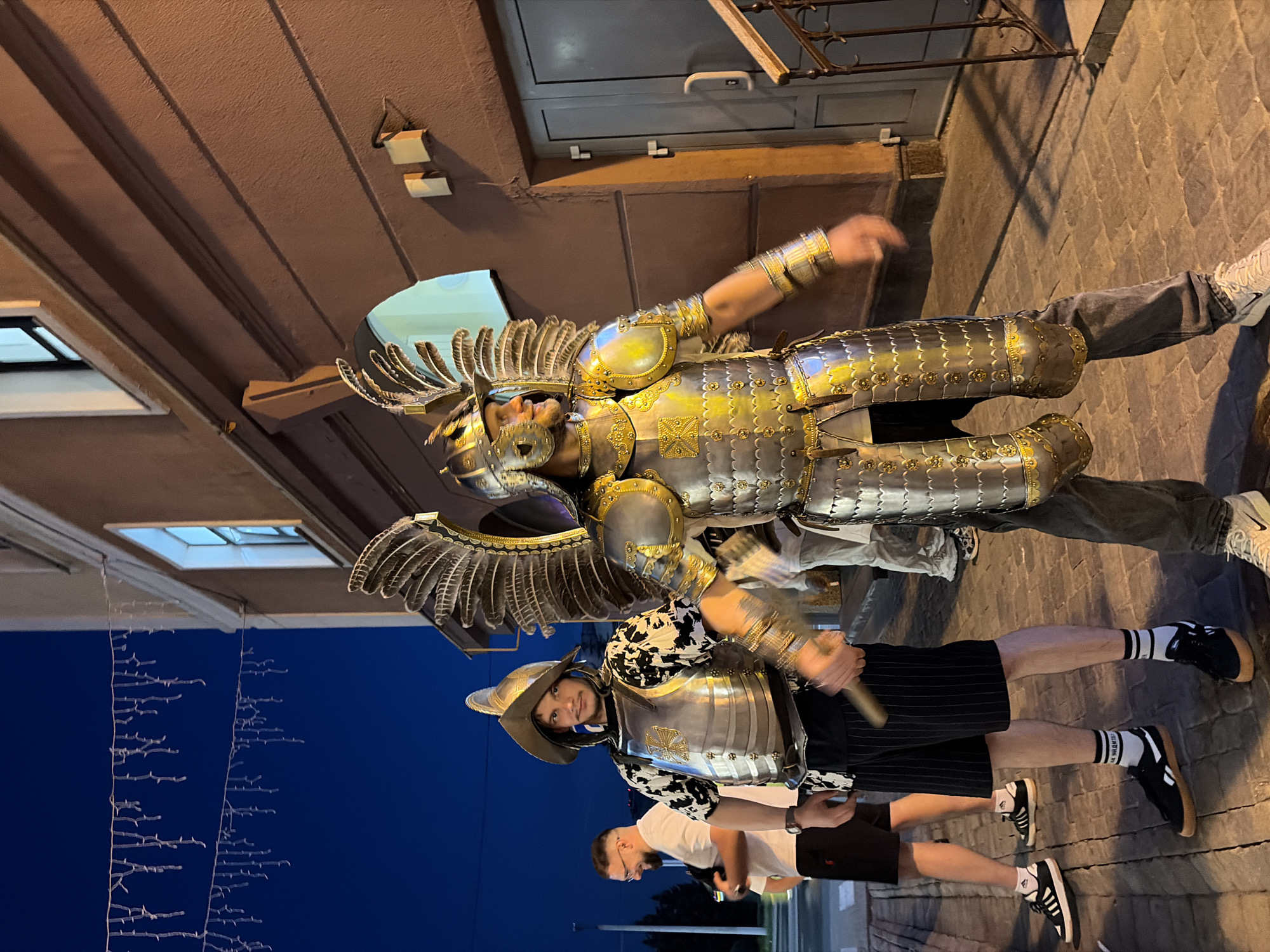 The image depicts two individuals dressed in stylized armor costumes standing on a brick-paved street, likely a pedestrian zone, based on the presence of other people casually dressed nearby. The upper figure is clad in full golden-colored armor with intricate details and a winged helmet. The lower figure is wearing partial silver-colored armor with a Spanish-style helmet. The background includes a building with a closed door and a window above. There are also festive string lights visible against a darkening sky, suggesting an evening setting. The location is Minsk, Belarus, based on the European architectural style, street pavement, and the presence of individuals dressed in specific styles.