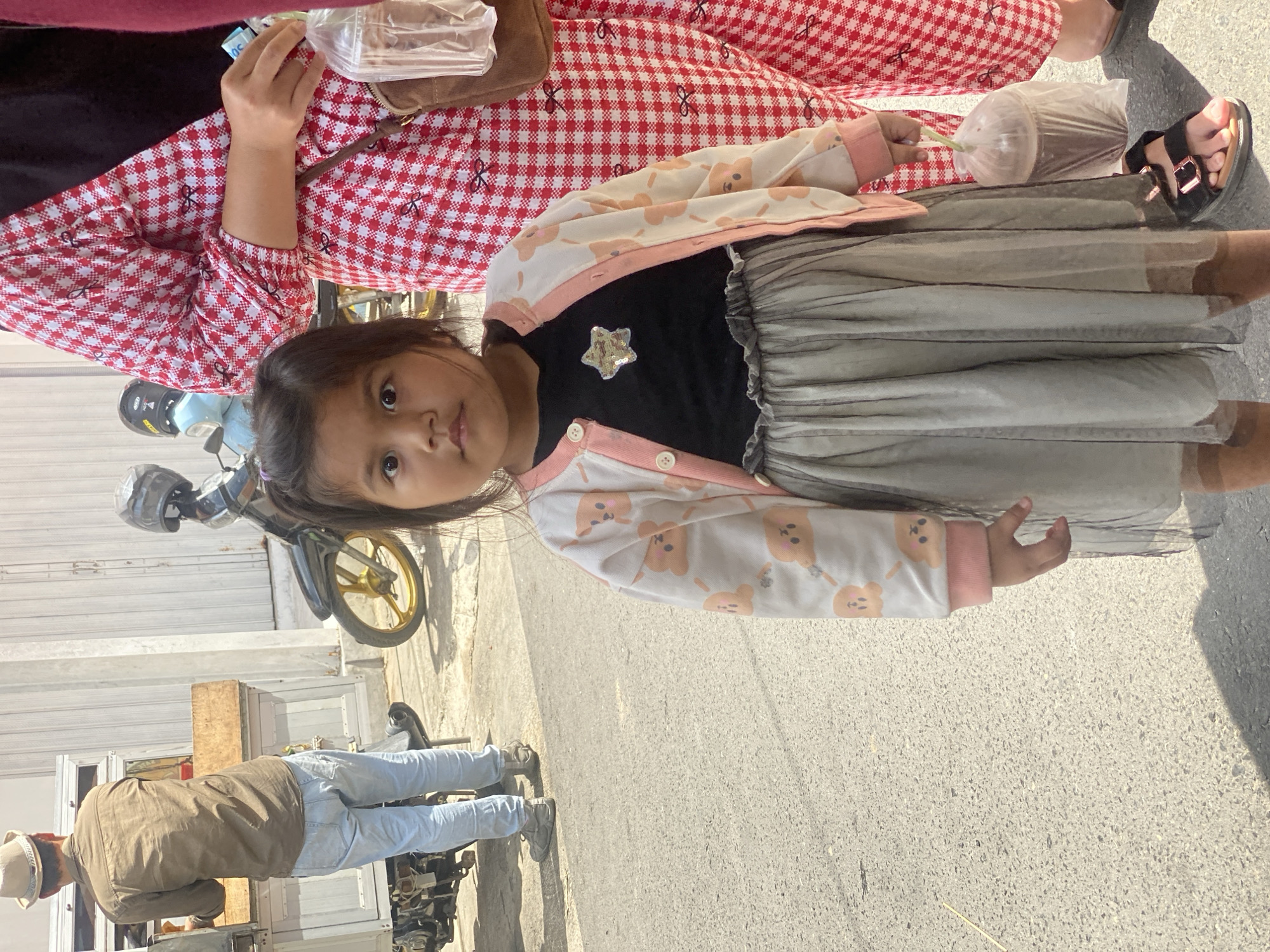 A young girl stands in the foreground on a paved street, looking directly at the camera. She wears a black shirt with a sparkly star patch, a light-colored cardigan adorned with cartoon animals, and a grey tulle skirt. In her right hand, she holds a plastic-wrapped pastry. To the right, partially visible, is a person wearing a red and white checkered garment. In the background, a man in a light brown shirt and light blue jeans stands behind a food cart. A motorcycle with gold wheels is parked nearby. The scene appears to be a street in Banjarbaru, Indonesia, likely during the daytime, as indicated by the lighting and shadows.