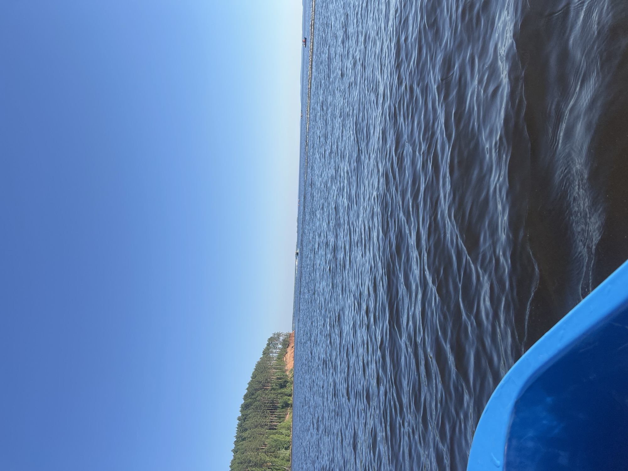 The image shows a view from a blue boat on the Kama River in the city of Kamskoe, Russia. The river is calm with small waves. On the left, a forested cliff overlooks the water. In the distance, several small boats are visible on the river. The sky is clear and blue.