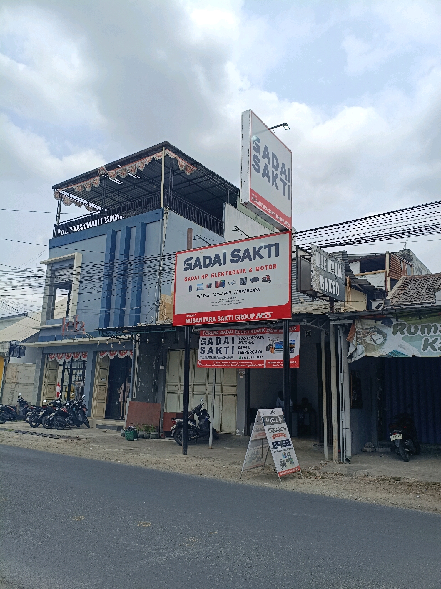 The image captures a street scene in Maguwoharjo, Indonesia, dominated by a multi-story building housing a business called "GADAI SAKTI," which appears to be a pawnshop specializing in electronics and motorcycles. The building has a blue and light blue facade with prominent signage. A large, vertical sign for "GADAI SAKTI" is prominently displayed on the right side of the building, featuring text and images of electronic devices and a motorcycle. Another A-frame sign for "GADAI SAKTI" is placed on the sidewalk in front of the building, displaying business hours and services. Several motorcycles are parked along the street in front of the shops. To the right of the "GADAI SAKTI" building, there is another business with a sign that reads "MILE AYAM BAKSO." The sky is overcast.