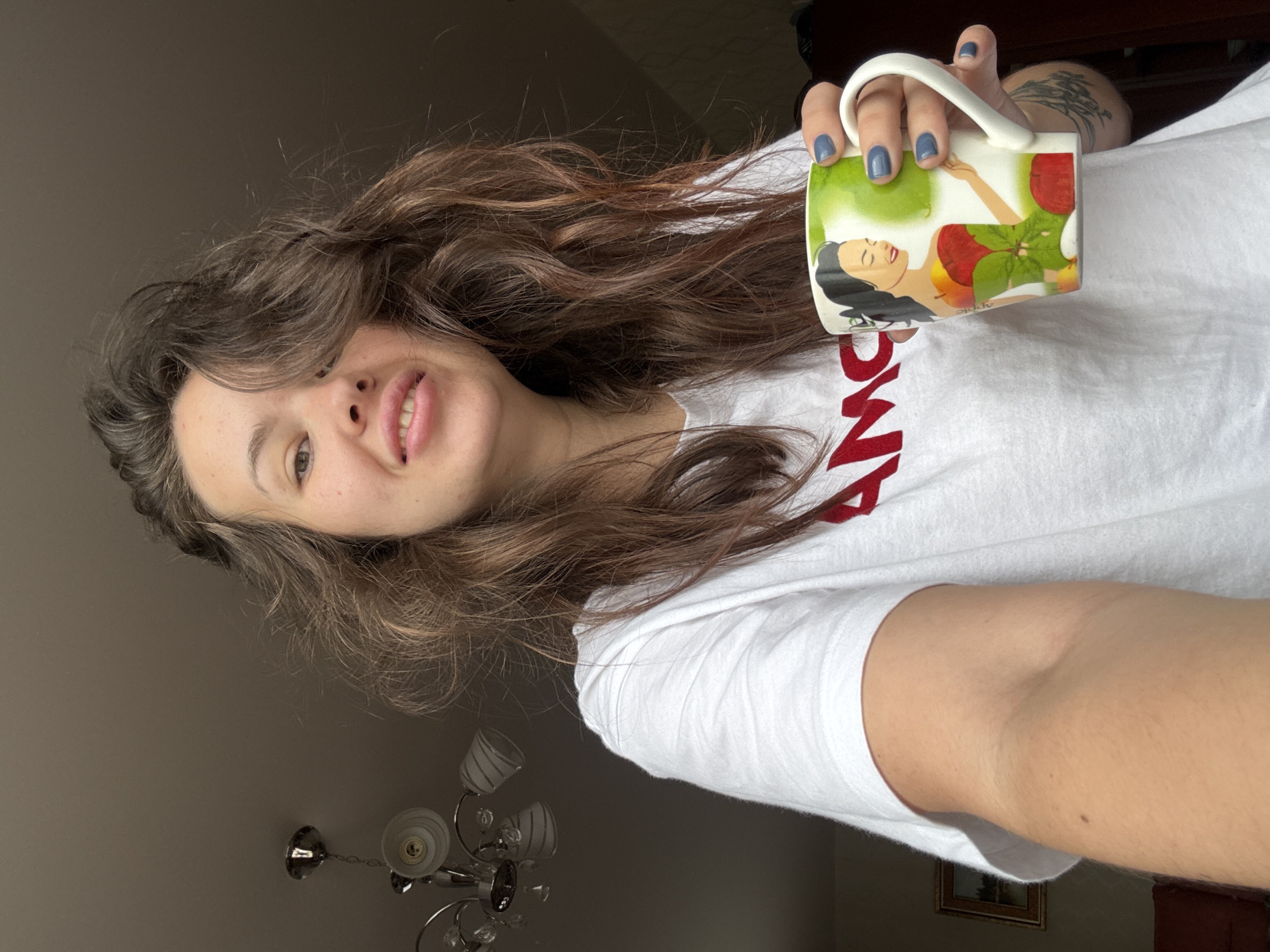 A woman with wavy brown hair is holding a mug with an illustration of a woman and apples. She is wearing a white t-shirt with red lettering. Her nails are painted blue. A chandelier is visible in the background, along with a beige wall and a framed picture. The location cannot be confirmed in Bely Yar, Russia with the information provided.