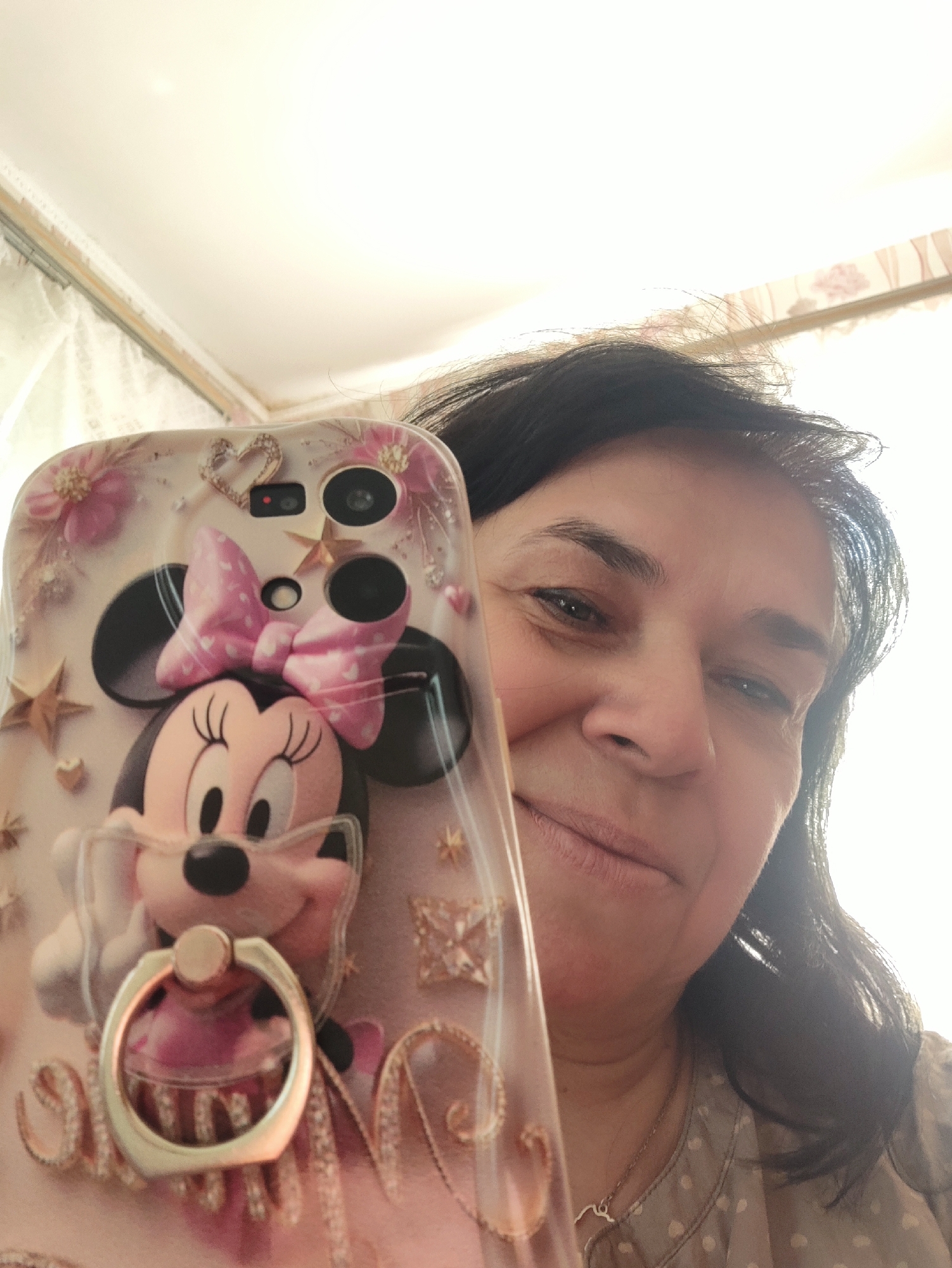 A woman is taking a selfie with a phone case decorated with Minnie Mouse. The woman has dark hair and is wearing a patterned shirt. The phone case has a pink bow and is adorned with small pink flowers, gold stars, and rhinestones. A ring holder is attached to the bottom of the case. The background appears to be an indoor setting with a white ceiling and patterned curtains.