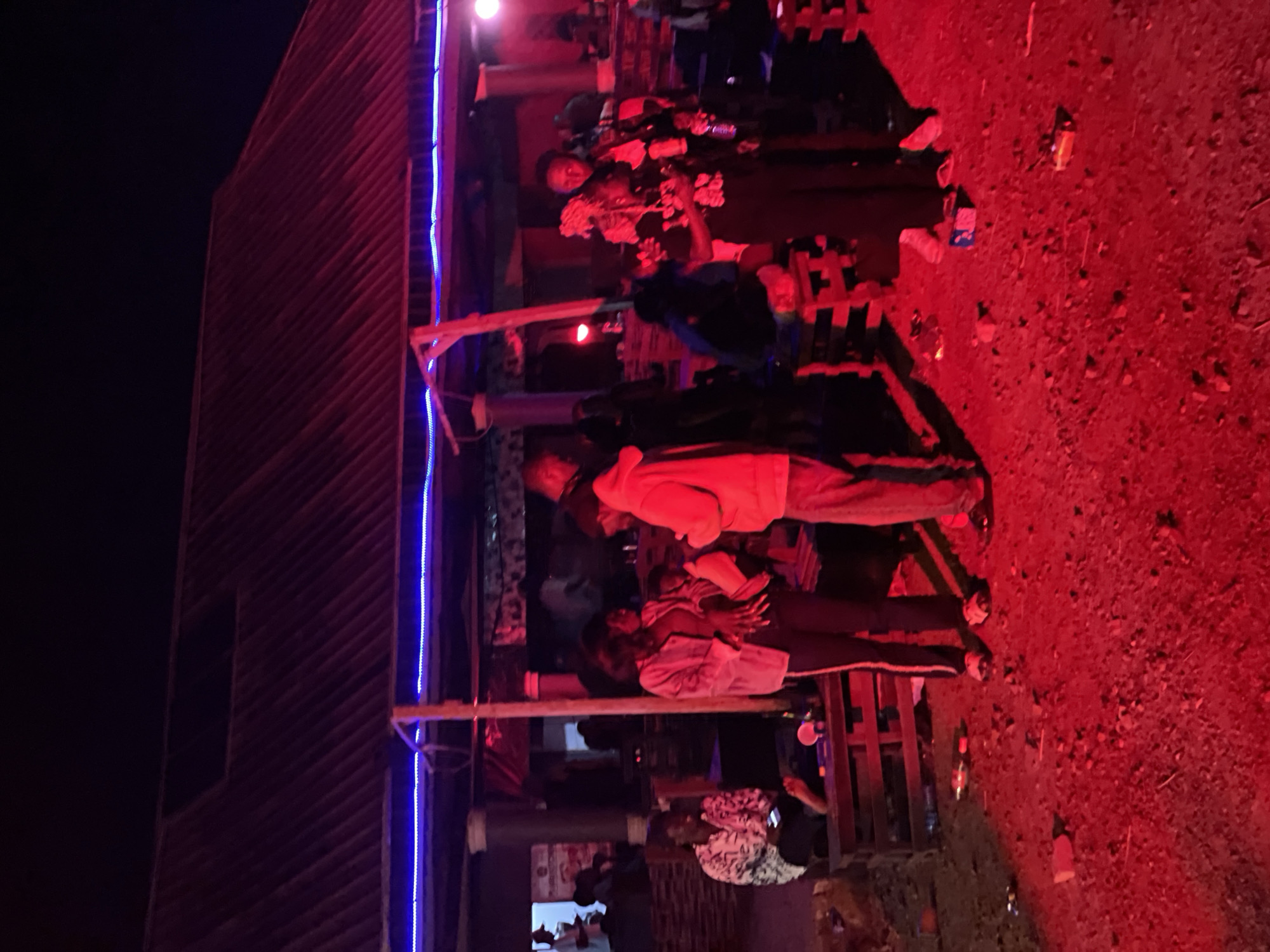 In Durumi, Nigeria, a group of people are gathered under a corrugated metal roof structure at night. The scene is illuminated by red and blue lights, casting a vibrant glow on the surroundings. In the foreground, a person in a patterned shirt sits on wooden pallets, looking at a mobile phone. To the right, several individuals stand around, some engaged in conversation. The ground is covered in dirt and debris, with scattered bottles and other objects visible. The structure appears to be an open-air establishment, possibly a bar or gathering place, with visible support beams and a dark ceiling. The overall atmosphere suggests a lively outdoor event.