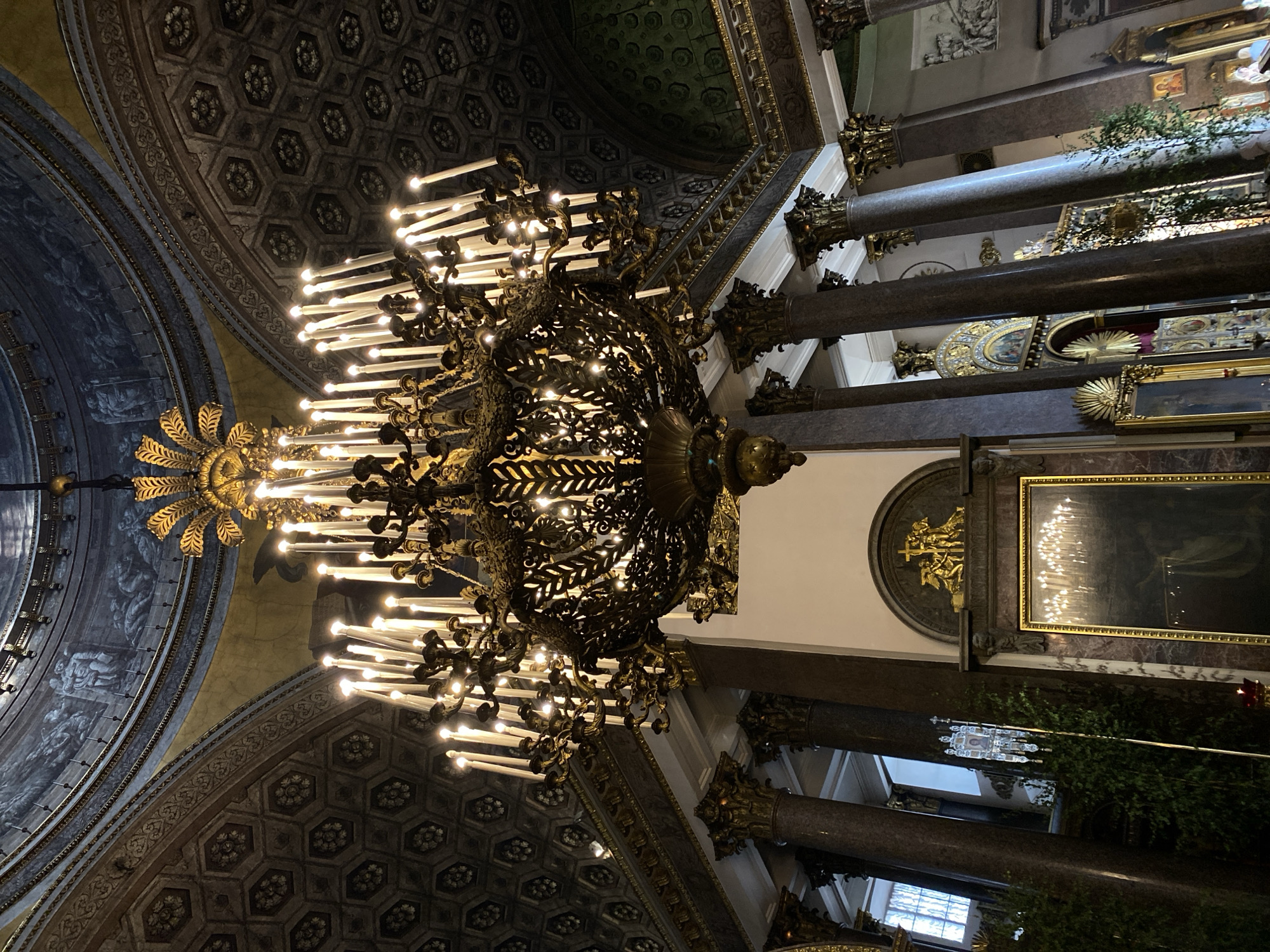 The image presents an opulent, upward-angled view inside a grand architectural space, likely a cathedral or palace, consistent with locations found in Saint Petersburg, Russia. The dominant features are several massive, ornate chandeliers casting a warm glow. The primary chandelier, centered and in the foreground, is dark-toned, possibly bronze or iron, intricately adorned with detailed, leafy metalwork and numerous candle-shaped light bulbs. Another golden, sunburst-like chandelier is partially visible to the left.
The ceiling and arches are richly decorated. A patterned vaulted ceiling features hexagonal coffers with dark floral or geometric motifs, bordered by elaborate gilded friezes. Curved arches are painted with frescoes depicting figures and scenes in a muted blue-grey palette, also outlined with gold trim.
Supporting the structure are substantial, polished stone columns, appearing dark grey or brown, possibly marble or granite, topped with highly decorative gilded Corinthian or composite capitals. The walls feature large, framed artworks; one prominent piece is a rectangular painting or icon with a golden frame, reflecting the chandelier lights. Above it, a semicircular niche holds golden relief sculptures, possibly depicting religious figures or narrative scenes. Greenery or decorative branches are interspersed among the columns in the background.
The lighting is predominantly artificial, originating from the many glowing chandeliers, suggesting either an evening setting or an interior where such illumination is constantly required. The overall atmosphere conveys grandeur, historical richness, and architectural magnificence. No discernible text is visible in the image.