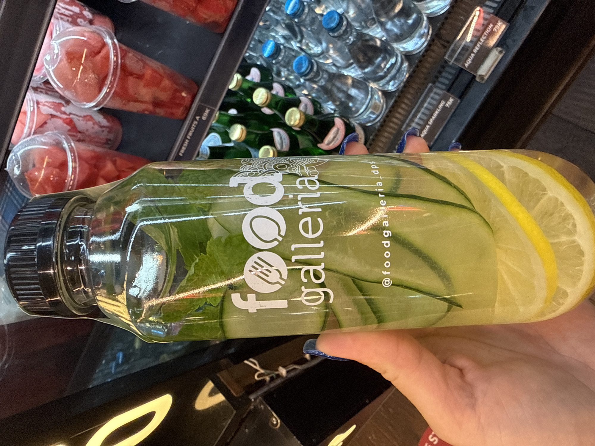 A hand, with blue-painted fingernails, holds a clear plastic bottle filled with cucumber-infused water and lemon slices. The bottle has a black cap and features a white logo that reads "food galleria" with a fork and spoon icon above it, and "@foodgalleria_dps" below. Behind the bottle, a refrigerated display case shows various items. In the upper background, several clear plastic cups are filled with chopped red fruit, likely watermelon. Below the fruit cups, green glass bottles with gold and pink caps are visible, followed by rows of clear glass bottles with blue caps. A price tag reading "AQUA SPARKKLING 75K" is visible to the right of the bottle. Another price tag, "AQUA REFLECTION 75K", is visible to the far right. The overall scene suggests a purchase from a store or cafe, possibly in Kuta, Indonesia, given the ".dps" in the Instagram handle, which often refers to Denpasar, the capital city of Bali, where Kuta is located.