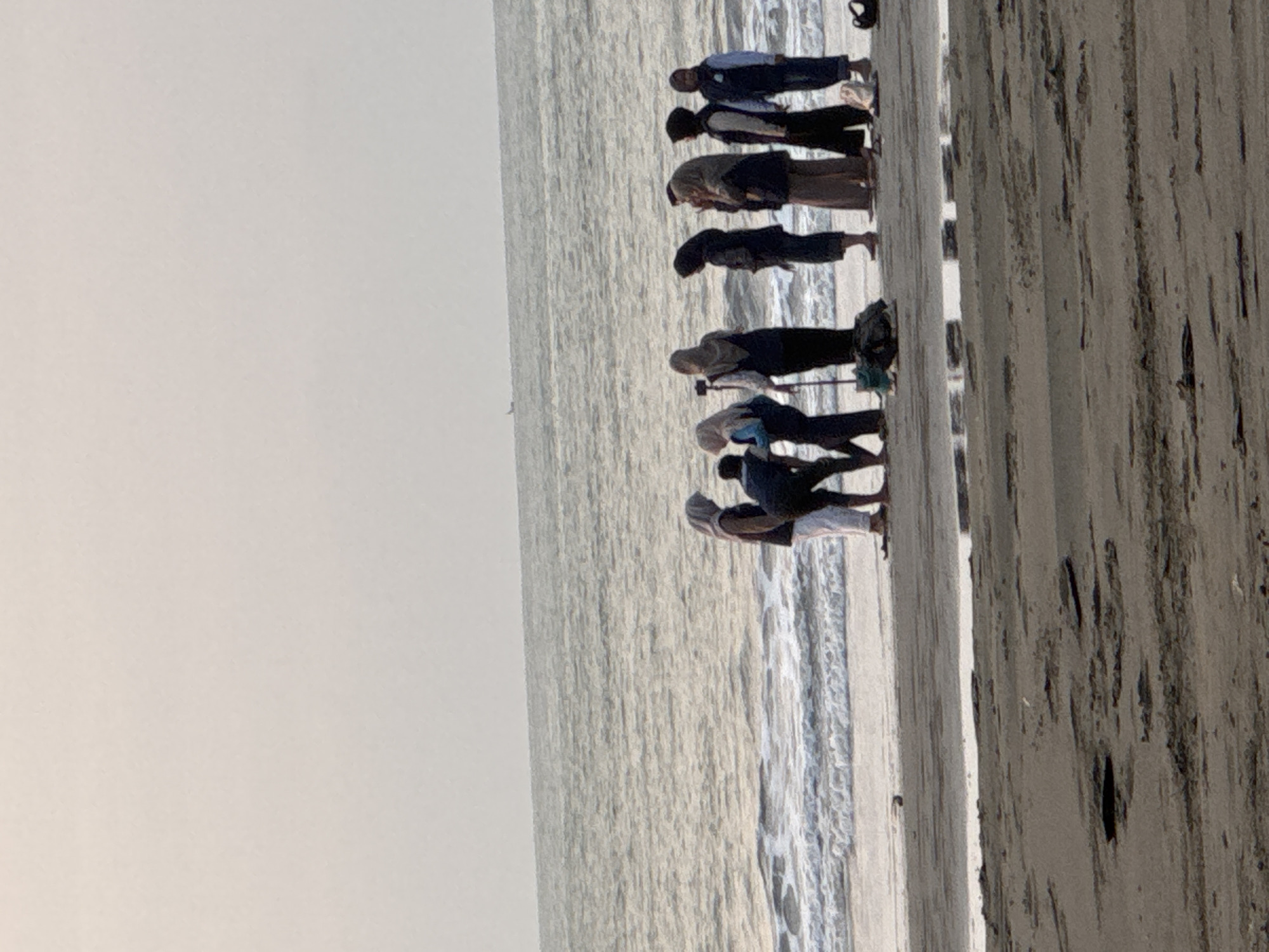 A group of seven individuals, six women and one man, stand on a sandy beach near the ocean. The women are dressed in hijabs and modest clothing. The man, wearing a dark shirt and light pants, is bent over. The group appears to be gathered near a selfie stick, suggesting they might be taking photos or videos. The ocean is visible behind them with gentle waves. The sky is overcast, and the overall lighting suggests it is either early morning or late afternoon. The beach is wet from the receding tide, with some debris visible on the sand. The location is identified as Reudeuep, Indonesia.