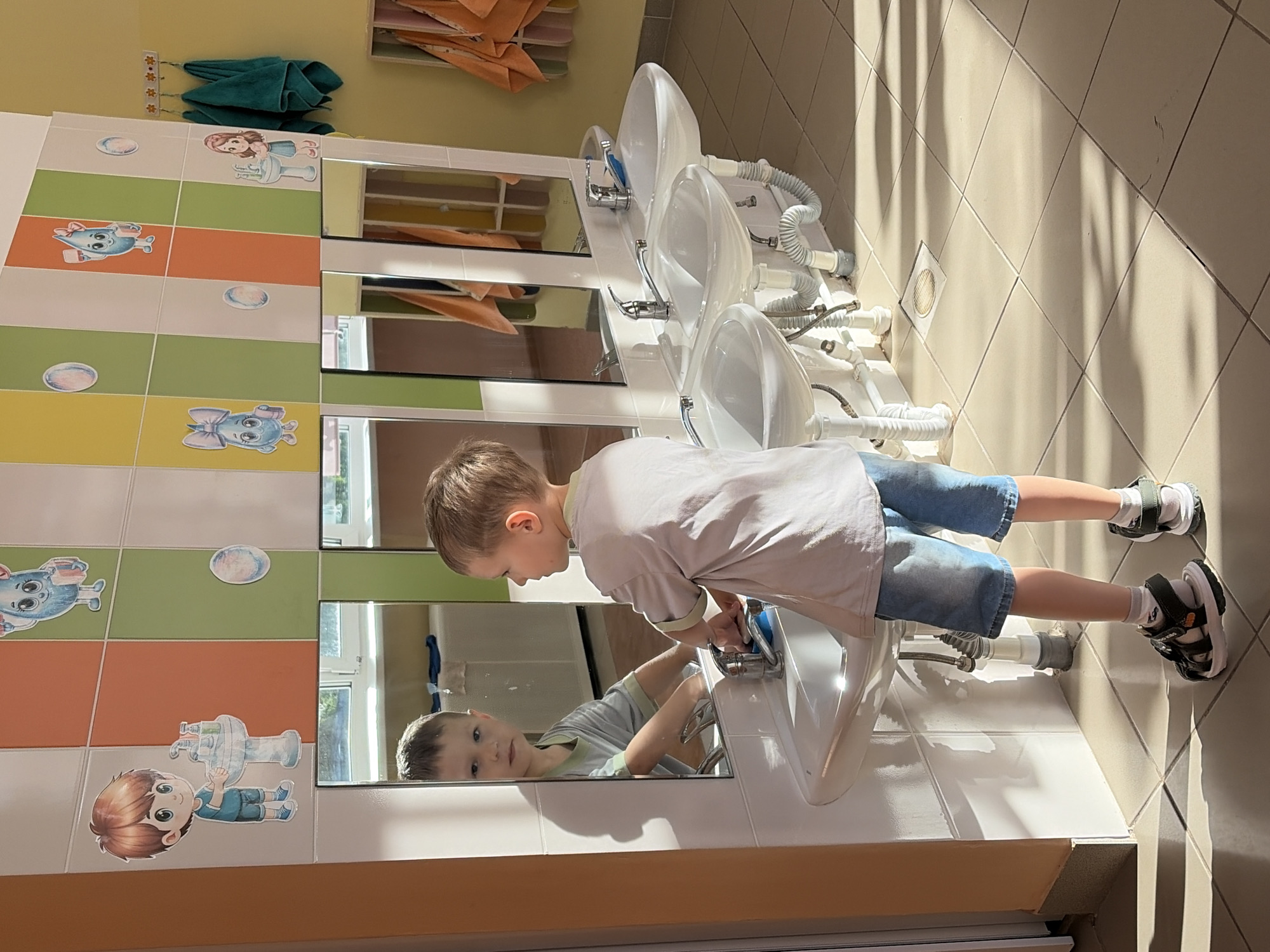 A boy is washing his hands in a children's bathroom in Chaikovsky, Russia. The bathroom has a row of three identical white sinks. The wall behind the sinks is decorated with colorful tiles featuring cartoon characters. Mirrors are hung above each sink. The boy is wearing a light-colored t-shirt, blue shorts, and sandals with socks. Sunlight streams into the room, casting shadows on the tiled floor. Towels are hung on a rack above the sinks.
