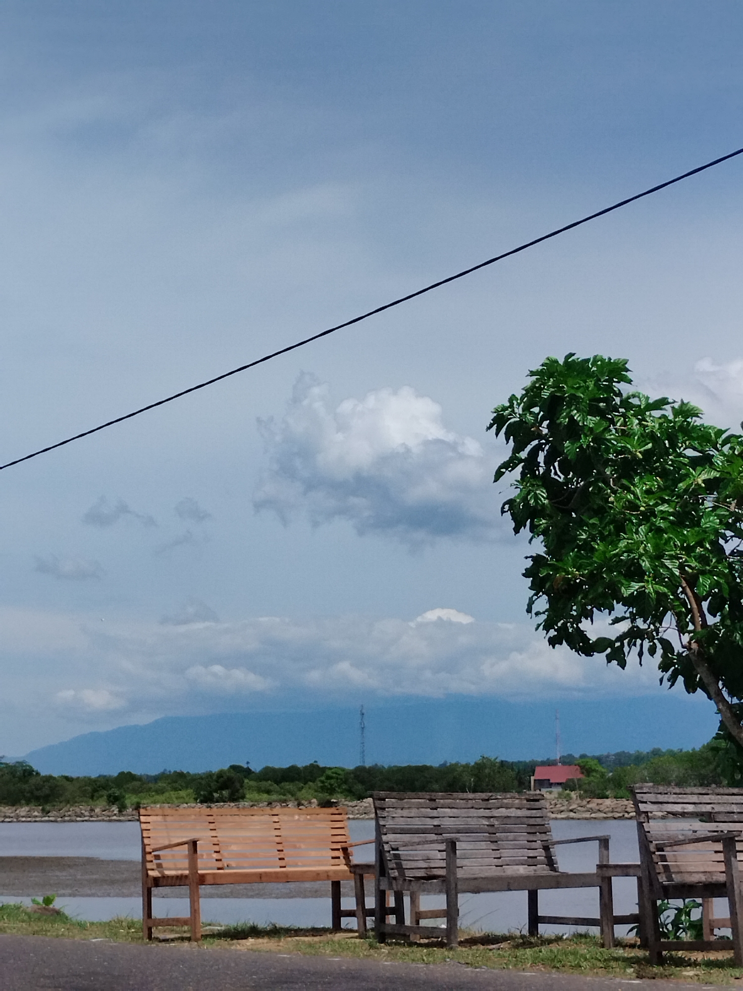 A tranquil waterside scene in Lhokseumawe, Indonesia, features two wooden benches facing a body of water. The sky above is a soft blue, dotted with scattered white clouds. A single black cable traverses the sky diagonally from the upper left to the upper right. To the right of the frame, a vibrant green tree with dense foliage stands tall. In the distance, a range of blue mountains is visible, with a red-roofed building nestled among trees in the mid-ground. The foreground consists of a paved road and a grassy verge.