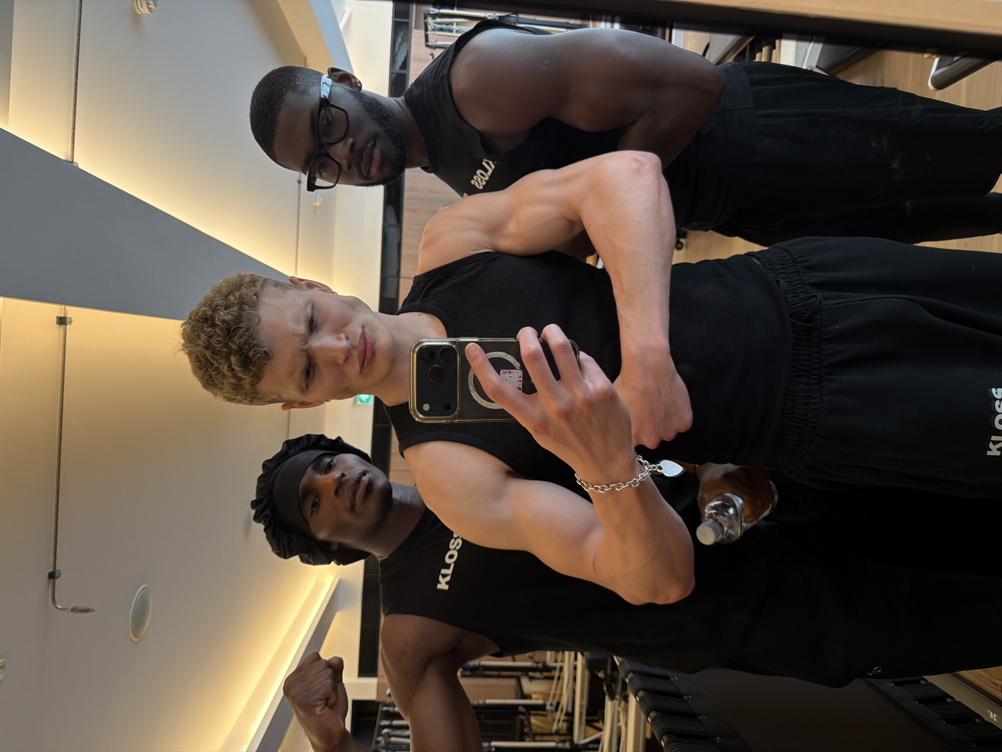 Three men are posing for a photograph in what appears to be a gym setting. The foreground is occupied by a fair-skinned man with short, curly blonde hair, holding a mobile phone in his left hand, seemingly taking a selfie. He's wearing a black tank top and black sweatpants. Behind him and to the left is a dark-skinned man with a black durag on his head. He is wearing a black tank top and posing by flexing his right bicep. Behind the fair-skinned man and to the right is another dark-skinned man wearing glasses, also wearing a black tank top. They are all in workout attire, specifically black tank tops and sweatpants with "KLOSS" printed on them. A water bottle is visible near the bottom of the frame, partially obscured by the man in the durag. In the background, gym equipment is faintly visible. The walls are clean and well lit, suggesting a modern and well-maintained facility. The gym is located in London, United Kingdom.