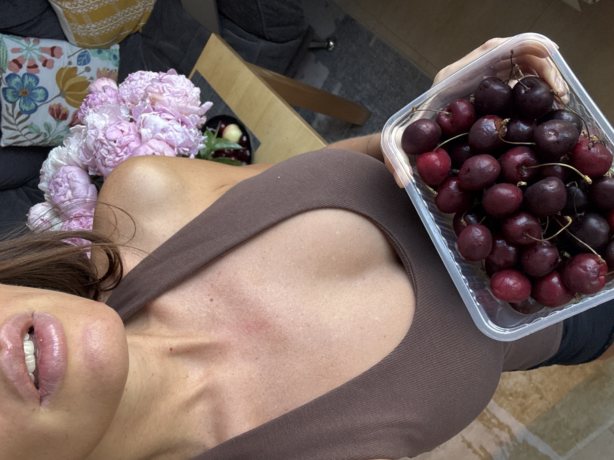 The image shows a close-up of a woman holding a container of cherries. She is wearing a brown tank top. In the background, there is a bouquet of pink peonies, a wooden table, and pillows on a couch. The location is not specified.