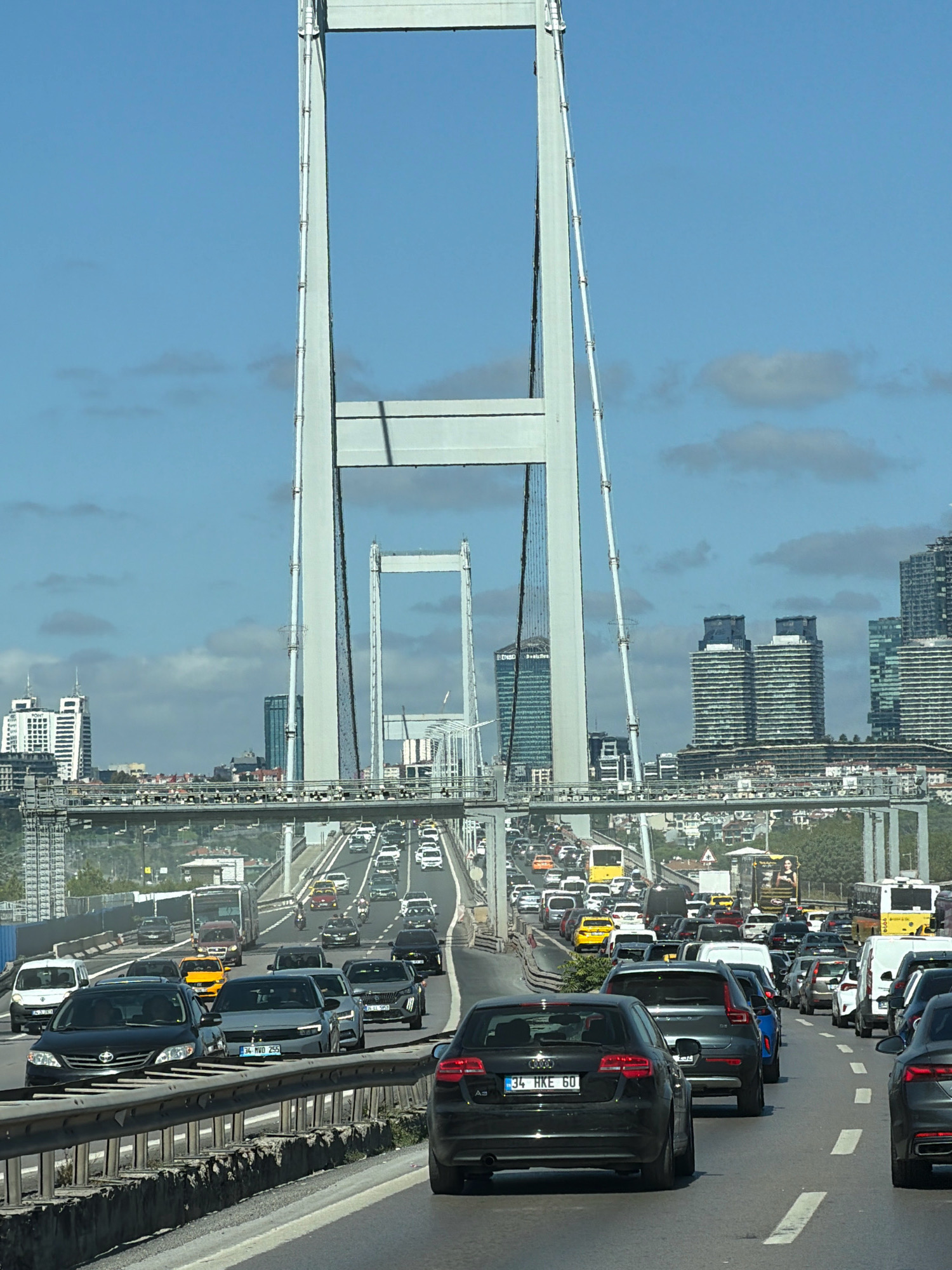 The image shows a busy highway with heavy traffic on a clear day. The foreground features several cars, including a black Audi A3 with license plate "34 HKE 60" and a grey sedan with license plate "34 MVO 255". In the background, a large suspension bridge dominates the skyline, with tall pylons reaching towards the blue sky. The bridge connects the city of Üsküdar, Turkey, to the other side of the Bosphorus strait. Several modern buildings are visible on the horizon, suggesting an urban environment. The highway appears to be elevated, with multiple lanes of traffic moving in both directions. A yellow bus is visible in the middle lanes, along with numerous other vehicles. The overall scene depicts a typical urban commute with significant congestion.