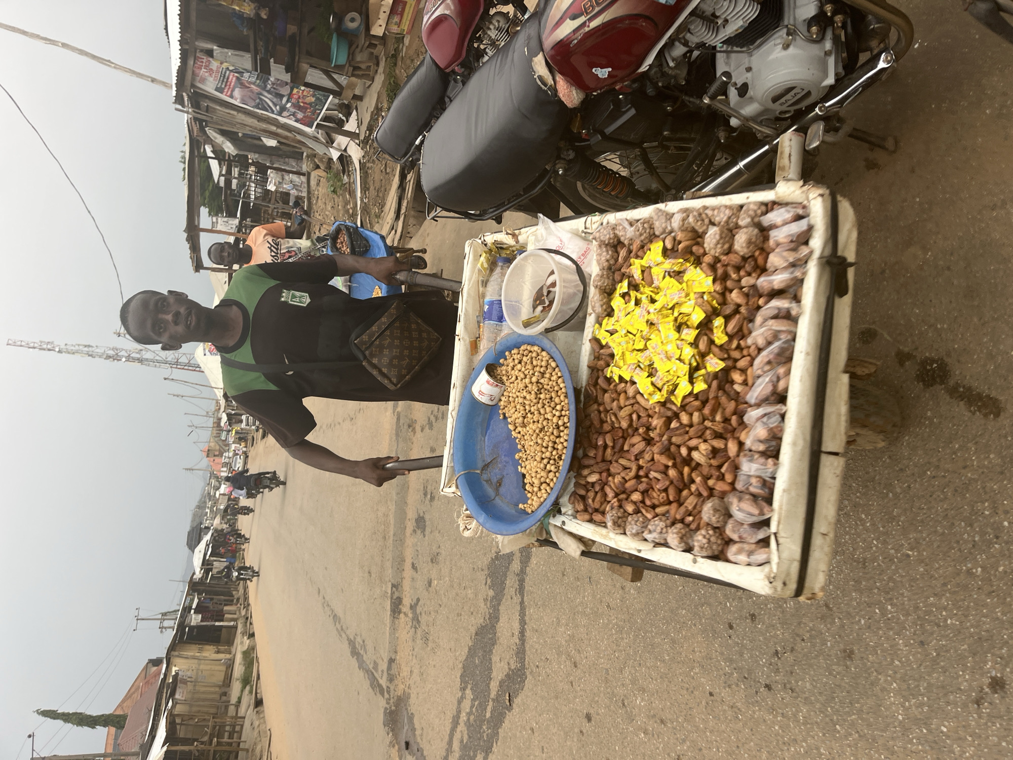 A man stands with a food cart on a street in Abuja, Nigeria. The cart is laden with various snacks, including nuts and dates. The man is wearing a black and green t-shirt and a Louis Vuitton-patterned bag. Behind him, another man stands near a stall. A red motorcycle is parked to the right of the food cart. The street is paved and shows signs of wear. Buildings line the street, some with corrugated metal roofs. The sky is overcast.