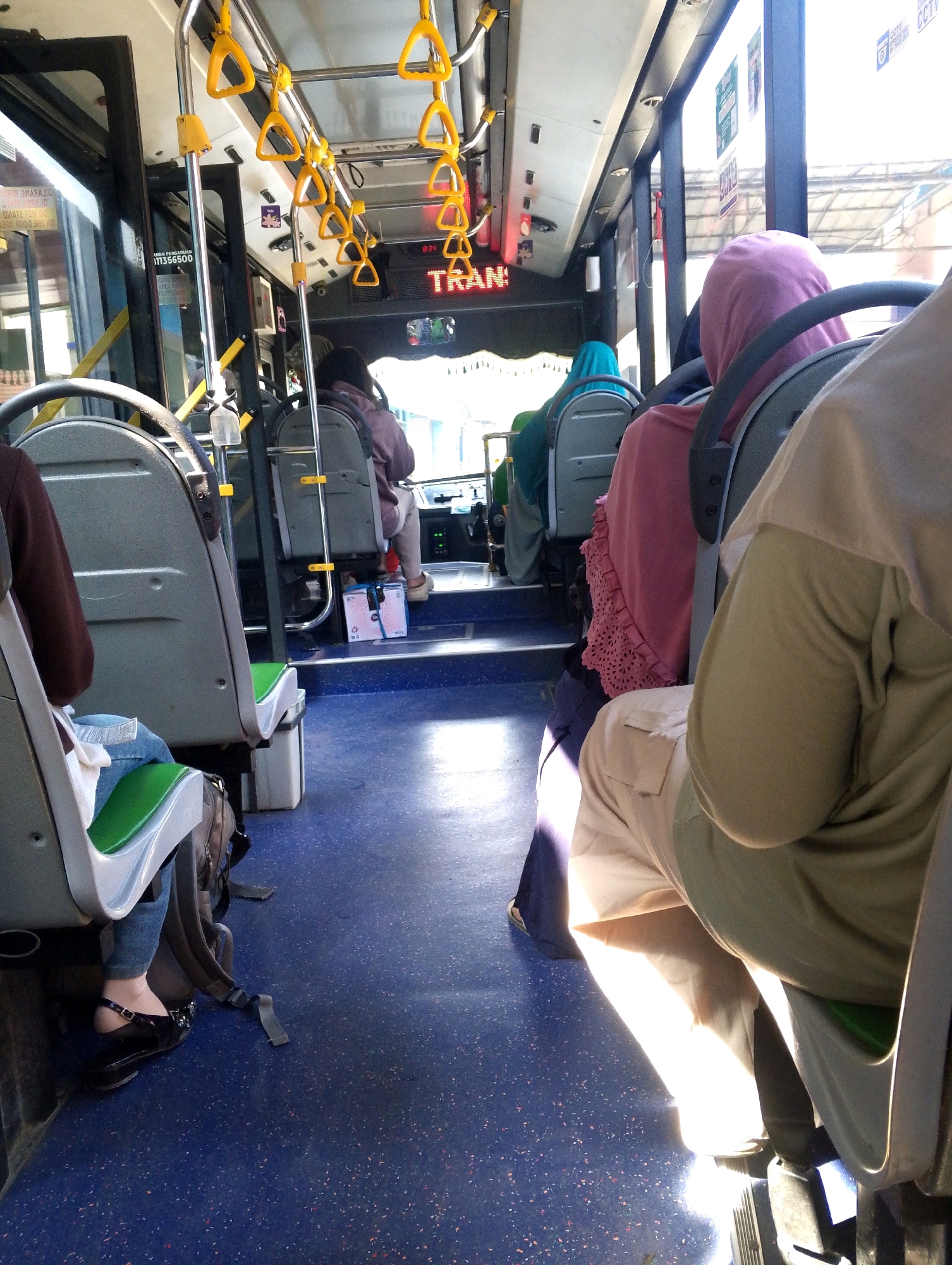 The image depicts the interior of a public bus with blue flooring and rows of gray seats. Several passengers are visible, mostly seen from the back and sides. Some passengers are wearing headscarves in various colors, including pink and green. The bus is equipped with yellow handrails hanging from the ceiling. Sunlight streams in through the windows, casting shadows on the floor. A digital display on the bus shows the word "TRAN" in red letters. Luggage is present, including backpacks and a pink and white bag on the floor near the front of the bus. The scene appears to be within a moving vehicle, suggested by the lighting and the angle of the shot.