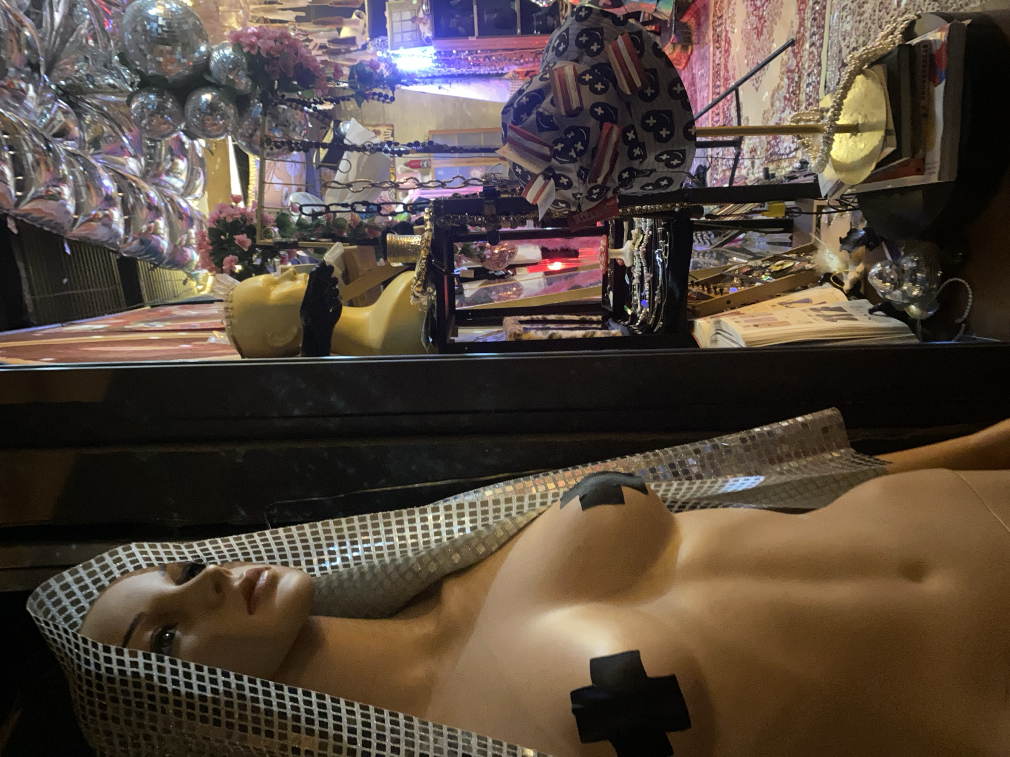 The image depicts a storefront display in Baan Nai Suan, Thailand. A mannequin, partially nude with black crosses covering the nipples and a silver checkered head covering, lies horizontally in the foreground. Behind the prone mannequin, there is a yellow mannequin head and a display rack containing jewelry, a hat with a star and stripe pattern, and various other items. In the background, there are reflective disco balls and floral arrangements. The overall scene appears cluttered and eclectic, typical of a retail environment.
