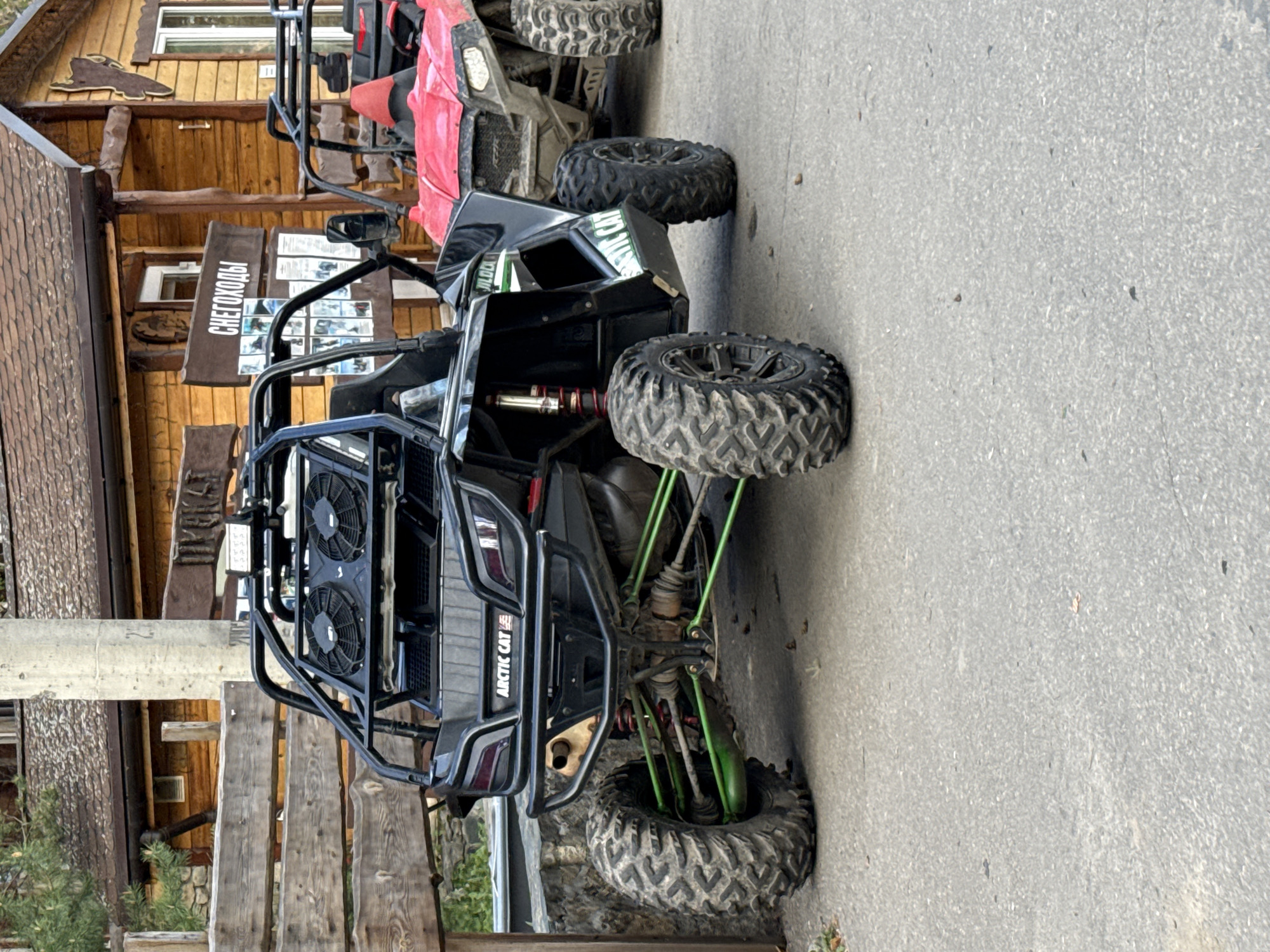Two off-road vehicles are parked in an outdoor setting. The vehicle closest to the viewer is black with green suspension components and the brand "ARCTIC CAT" visible. Partially visible behind it is a red off-road vehicle. Behind the vehicles is a wooden structure featuring a sign in Russian, "СНЕГОХОДЫ", which translates to "Snowmobiles". This is consistent with the location being Zolotoi Plyazh, Russia. The ground appears to be paved with asphalt. There are no people visible in the image.