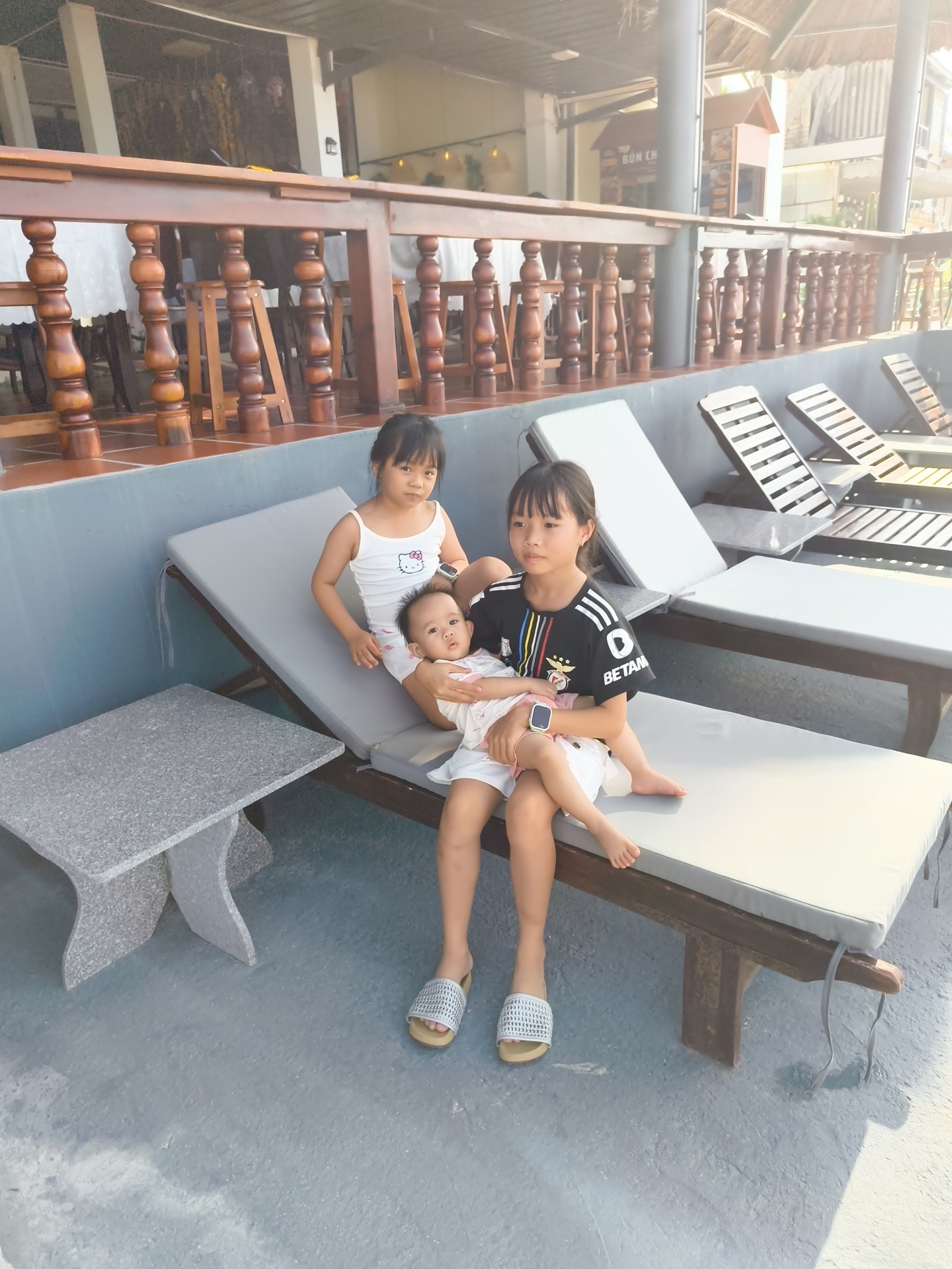 Three young females are posing for a photograph on lounge chairs in what appears to be an outdoor seating area of a restaurant in Phu Thuy Ward, Vietnam. The youngest, a baby, is held in the arms of the middle female, who is wearing a black and white t-shirt. The oldest female, wearing a white tank top, is seated next to them. A stone table is to the left of the lounge chairs. The background features wooden railings, tables, and chairs, suggesting the location is an eatery.