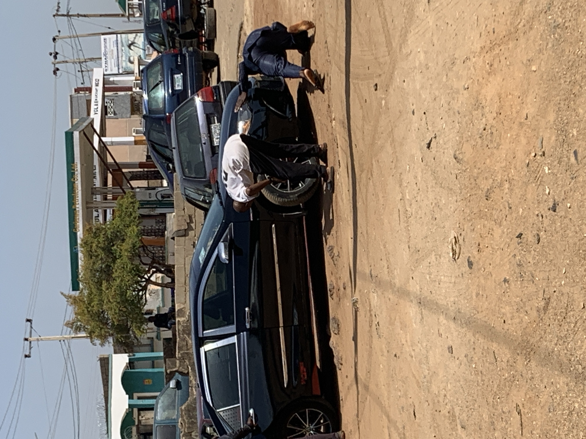 The image shows a street scene in Jos, Nigeria. The ground is dirt and gravel. A black sedan is parked alongside other vehicles. Two individuals are interacting with the sedan: one in a white shirt appears to be working on the front driver's side tire, while another person in dark clothing is on the hood of the car. A gas station is visible in the background with green and white branding. Utility poles and wires line the side of the road. Buildings and a tree are visible in the background.