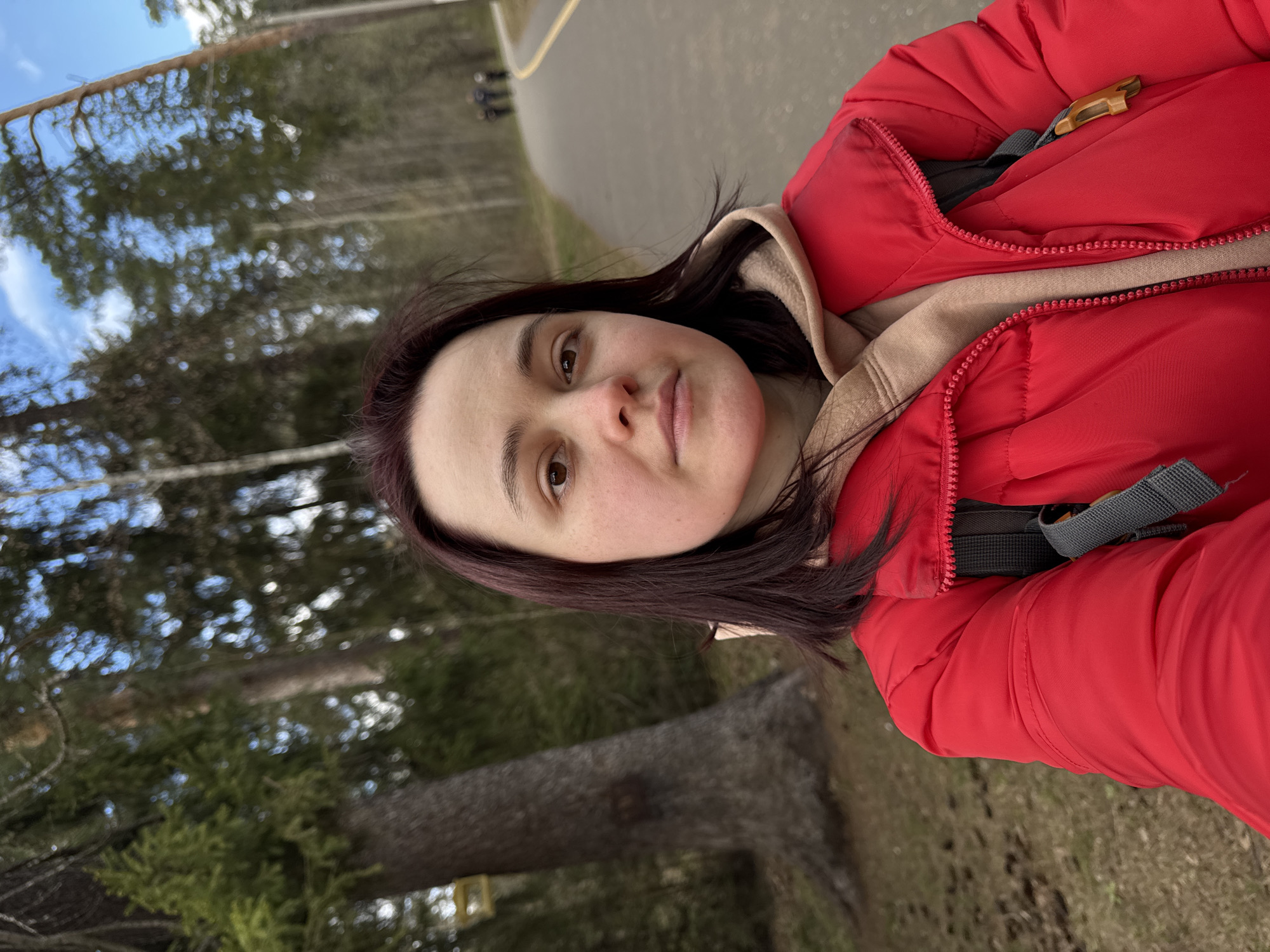 A woman in a red jacket and beige hoodie takes a selfie on a path in a wooded area. The path appears to be paved, with a yellow line painted on it, and some people are walking in the distance. Trees and foliage are visible on either side of the path. The sky is partly cloudy. The location appears to be a park. I cannot confidently identify a specific location in Izhevsk, Russia without additional information.