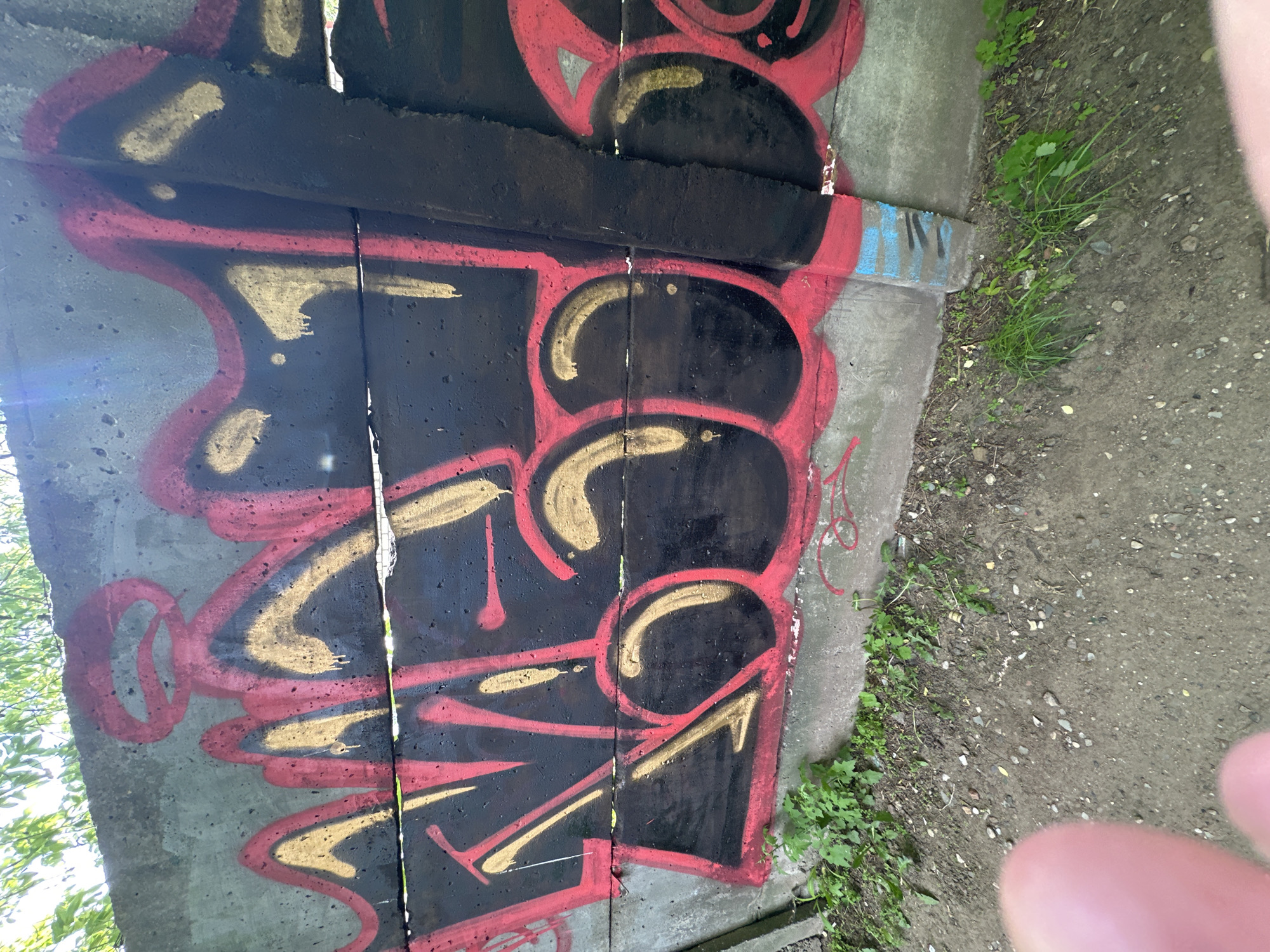 The image depicts graffiti on a concrete wall. The graffiti is characterized by bold black lettering outlined in red and accented with gold. The letters appear stylized and abstract. The wall is weathered, with visible cracks and signs of age. Adjacent to the wall is a patch of overgrown grass and dirt. The photo was taken during the daytime, and there is evidence of bright sunlight. There is a part of the photographer's hand in the lower right corner. The location is Minsk, Belarus, based on the provided information.