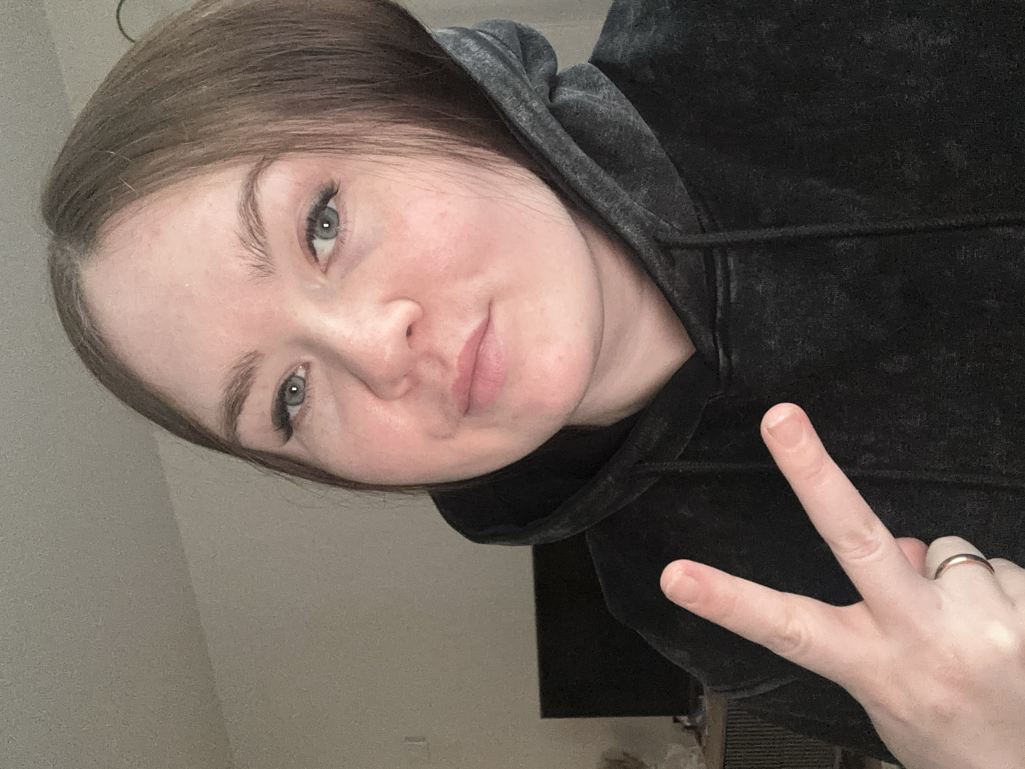 A young woman is captured in a close-up, slightly tilted selfie, likely taken indoors in Chaikovsky, Russia. She has light brown hair, which is parted on the side and falls over her forehead. Her blue-green eyes are prominent, and she has visible eyelashes. Her skin appears fair, with some freckles or natural marks visible on her cheek and forehead. She is wearing a dark, possibly charcoal grey or black, textured hoodie with a hood pulled up and resting on her shoulders. Her lips are lightly colored, and she has a calm expression with a subtle hint of a smile.
Her right hand is raised, making a peace sign (V-sign) with her index and middle fingers extended. A simple gold-colored band ring is visible on her ring finger. The background is simple and out of focus, appearing to be a light-colored wall, indicating an indoor setting. There is no visible text in the image. The lighting suggests a well-lit interior, though the specific time of day is not discernible. The overall mood is casual and relaxed.