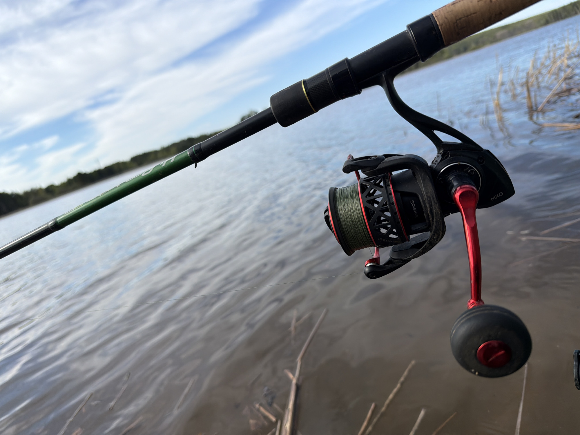 A fishing rod and reel are positioned in front of a body of water. The rod is angled from the upper left to the center of the frame, with the reel prominently displayed. The reel is black with red accents and green line. The handle of the reel is red with a black knob. Behind the rod and reel, the body of water extends to the horizon, with vegetation along the shoreline. The sky is visible in the background with scattered clouds. The location is unspecified. The city of Chaikovsky, Russia is not confirmed.