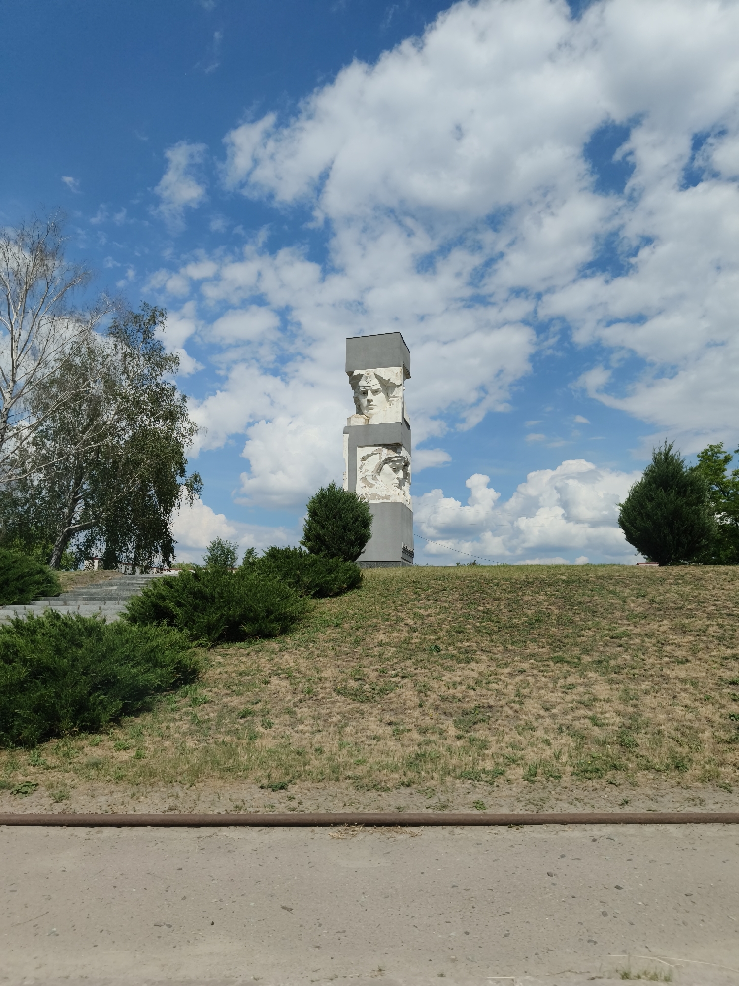 This media file depicts a monument set against a bright blue sky with scattered white clouds, likely located in the Krynychanska aglomeration, Ukraine.
The central subject is a tall, grey, rectangular monument positioned on a grassy hill. It features two prominent bas-reliefs of human faces on its visible side; the upper one shows a person, possibly a woman, wearing a star-emblazoned headpiece, while the lower one depicts another stylized face. The monument's design suggests a commemorative or heroic theme, typical of Soviet-era memorials.
To the left of the monument, a mix of trees is visible: one with bare, slender branches, resembling a birch, and another with dense green foliage. Lush green bushes are arranged around the base of the monument and line the slope of the hill, some in dense clusters. The hill itself is covered in sparse, dry-looking grass and patches of earth, indicating a warm, possibly dry, season.
In the immediate foreground, a concrete path or road stretches horizontally across the bottom of the frame. Above this path, a rusty, horizontal pipe or rail runs along the base of the grassy incline.
The sky is a vibrant blue, filled with fluffy, white cumulus clouds, suggesting clear and sunny weather conditions during daytime. There are no people visible, and no specific activity or event is taking place, presenting a static landscape scene. No discernible text is visible on the monument or elsewhere in the image.