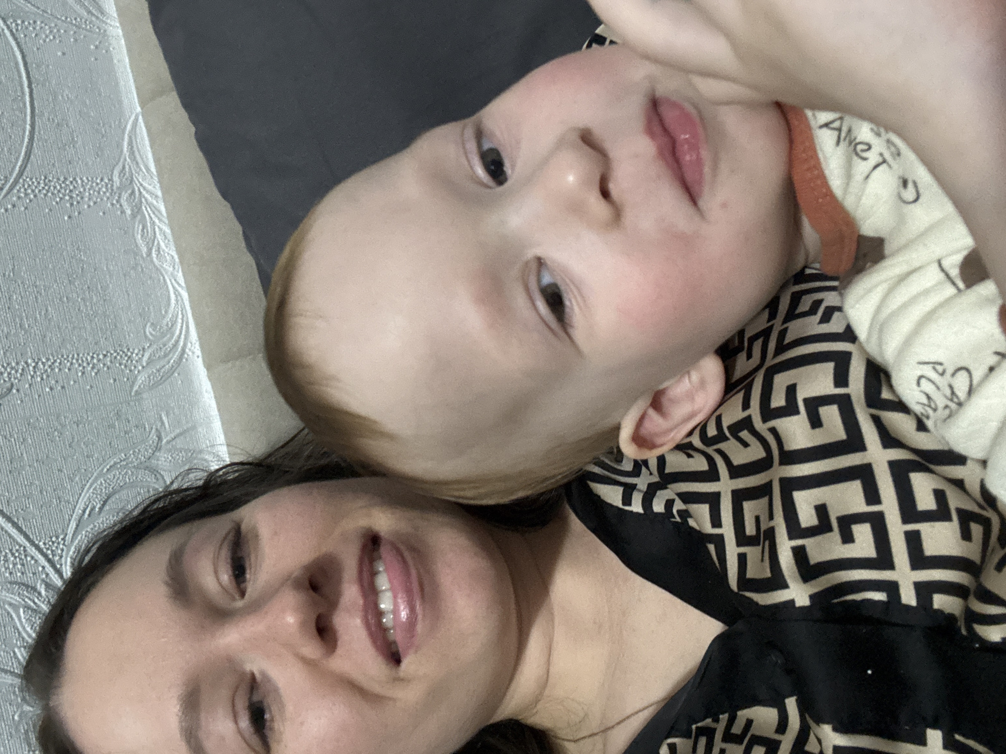 A photograph depicts a woman and a child lying down, possibly on a bed or other soft surface. The woman is at the bottom left of the frame, smiling, while the child is positioned above her. The child appears to be looking directly at the camera. The woman has dark hair, and the child has lighter hair. The child is wearing clothing with lettering on it. A black and beige patterned piece of fabric is visible. The environment includes a patterned wall or fabric in the background. There is no information in the image that indicates the precise location in the city of Ферма, Russia.