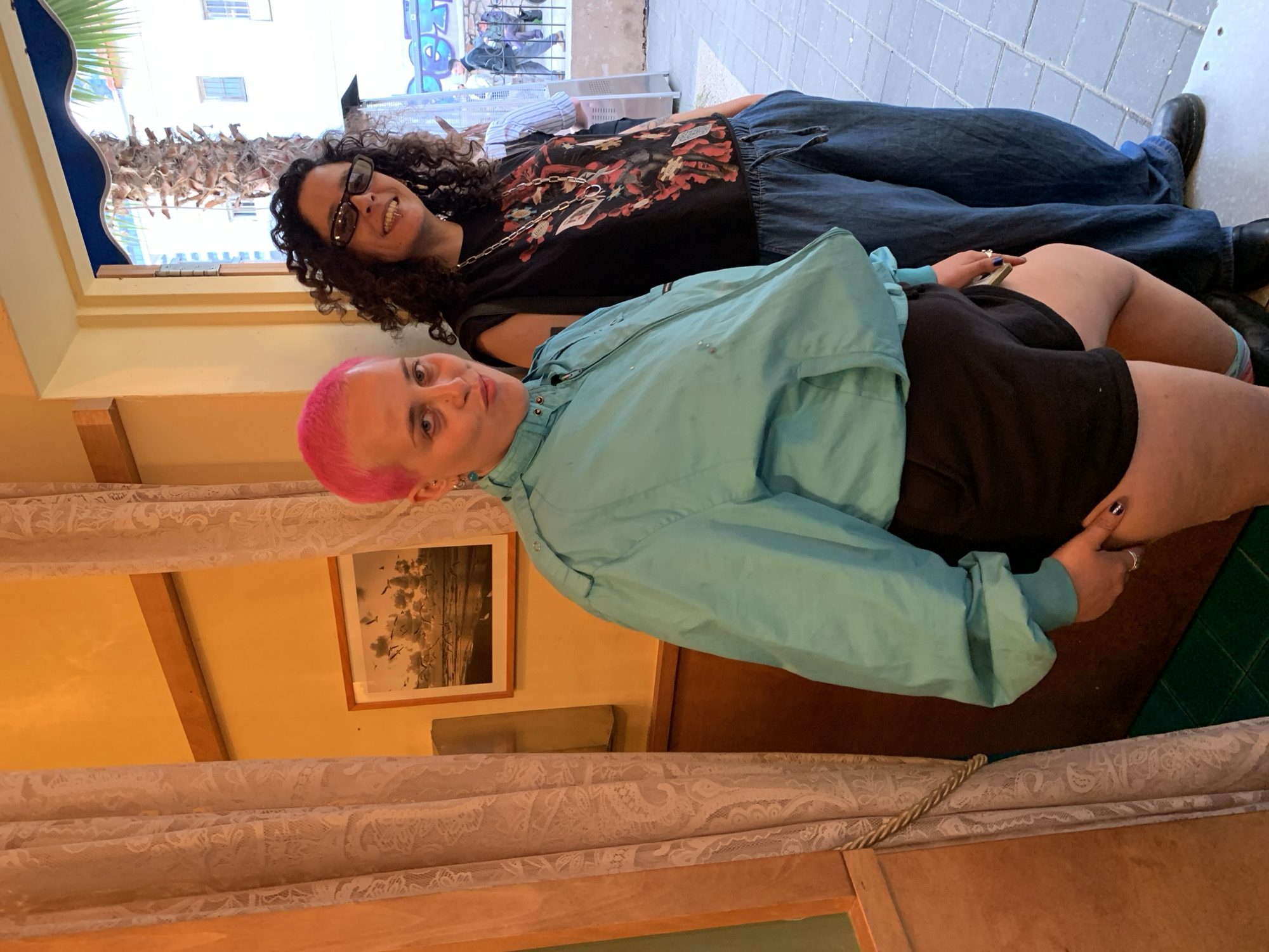 Two individuals are captured in what appears to be an indoor setting in Tel-Aviv, Israel.
The first person, with short, bright pink hair, is wearing a light blue windbreaker jacket over a black top and shorts. This person is positioned towards the front of the frame, with the focus on their face, which is turned towards the camera.
Behind this individual, another person with dark curly hair and glasses is visible. They are wearing a black shirt with a graphic design and blue jeans. This person is smiling and is lying down in a way that suggests they are posing for the photo along with the person in front.
Both individuals seem to be in a relaxed state.
The setting includes a window with a partial view of a building across the street. The interior is decorated with a framed picture on the wall. A lace curtain is visible in the foreground. The overall lighting appears to be a mix of natural light coming through the window and artificial lighting within the room.