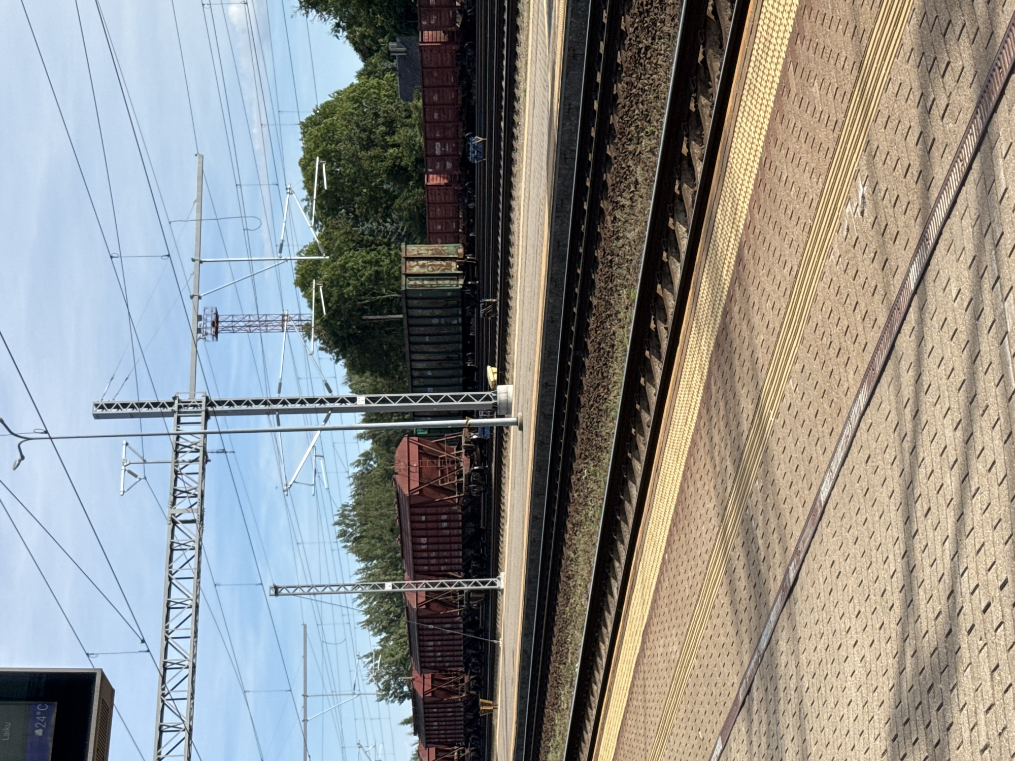 This is a photograph of a train station platform with multiple train cars visible on tracks in the background. The platform has a textured surface with tactile paving in yellow and grey. The train cars are primarily red hopper cars, with one green gondola car visible. Overhead power lines and supporting structures are prominent in the sky. Trees line the background beyond the train tracks. The image appears to be taken on a sunny day with a clear blue sky. The scene is located at the Šiauliai railway station in Šiauliai, Lithuania.