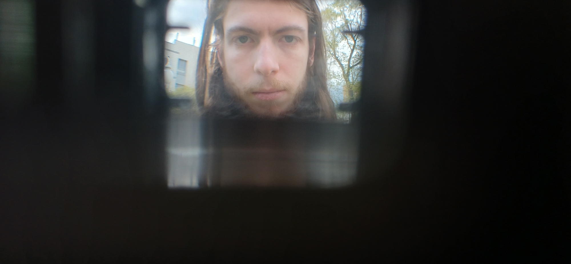 A person's face is seen through a small rectangular opening, likely a peephole. The person has long brown hair and fair skin. Outside, a building and trees are visible, suggesting an urban environment. The location is Unknown, Unknown.