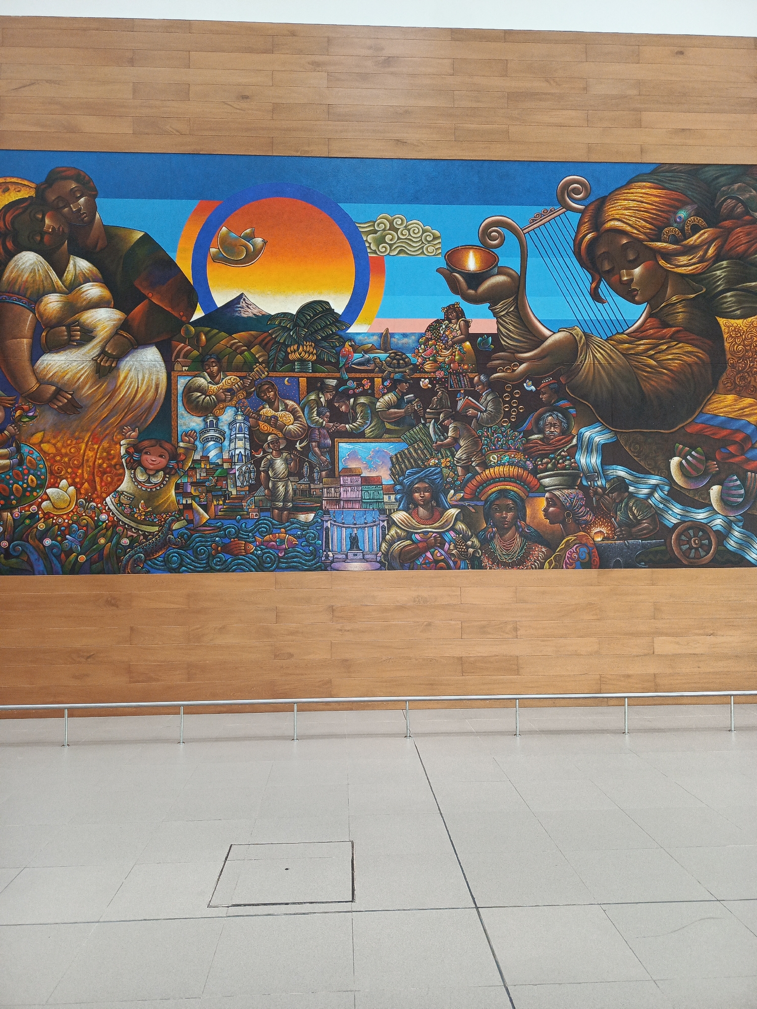 The image shows a large mural painted on a wall, depicting a vibrant and detailed scene of community and cultural life. The mural is set against a backdrop of light brown wooden planks. In the foreground, a polished concrete floor with visible grout lines and a rectangular access panel is visible, along with a silver railing supported by metal posts.
The mural itself is a complex narrative tapestry. On the left, a couple embraces, with the woman appearing pregnant, and they are positioned against a large, abstract sun with a mountain peak and a bird in the sky. To the right of this couple, a multitude of figures and elements are depicted. There are people engaged in various activities, including playing music, working, and socializing. Many individuals are dressed in colorful, traditional attire, with elaborate headdresses and jewelry. A prominent figure on the right side is a woman with flowing golden hair, playing a harp-like instrument, with a lit candle in her outstretched hand. Below her, various scenes unfold, showing buildings resembling a city with a lighthouse, boats on water with fish, and more people in different occupations. The overall impression is one of abundance, cultural heritage, and the interconnectedness of life.
This mural is located in the terminal of the José Joaquín de Olmedo International Airport in Guayaquil, Ecuador.
