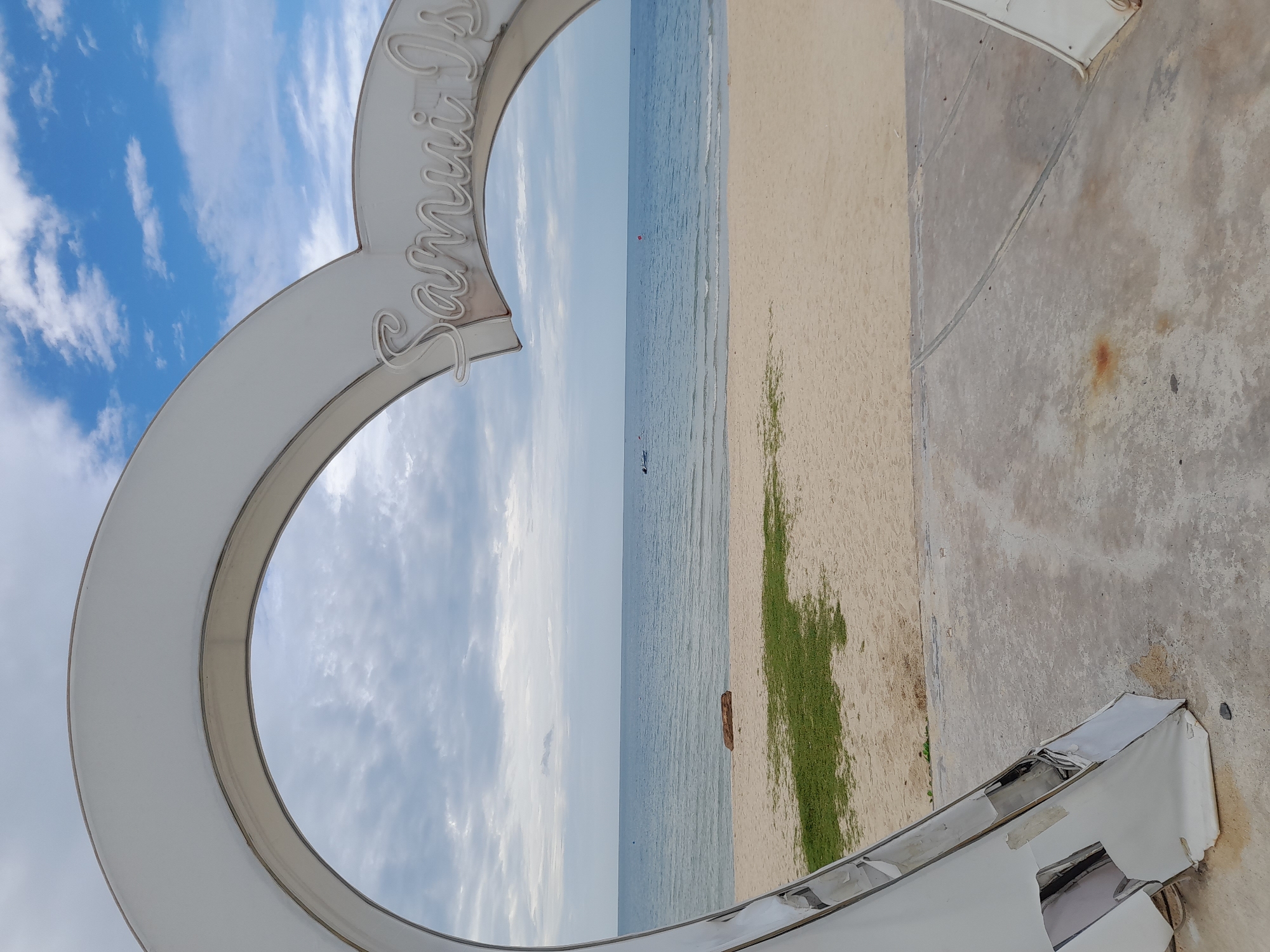 A heart-shaped sculpture frames a view of a beach and the ocean. The sculpture is white and appears to be made of metal and plastic. It has the words "Samui Is" written in script on its upper right arc. The beach is sandy with patches of green grass. The ocean is blue and calm, with a few small boats visible in the distance. The sky is partly cloudy. The foreground shows a concrete surface with some debris.
