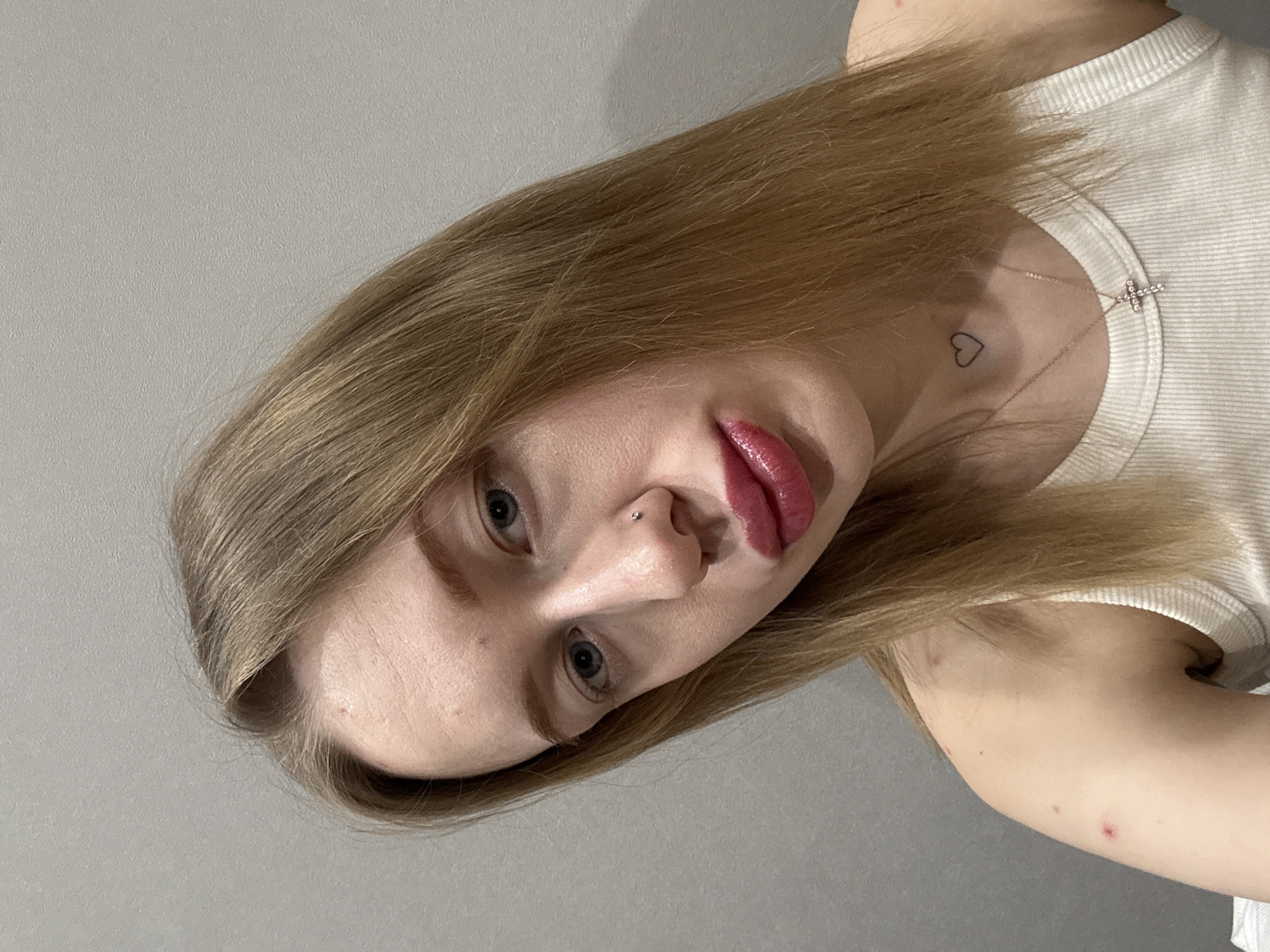 A young adult female is depicted in a close-up portrait, facing the camera with a slight head tilt to her right. Her hair is long, straight, and light brown/blonde, falling over her shoulders. She has blue eyes, a silver stud piercing on her right nostril, and her lips are pursed, coated with glossy, dark pink lipstick. Visible on the left side of her neck is a small, outline heart tattoo. She wears a thin, rose gold-colored chain necklace with a small, jeweled cross pendant and a white ribbed tank top. Several small red blemishes are present on her left upper arm/shoulder area. The background is a plain, light gray textured wall, indicating an indoor setting. The specific location within the city of Rostov-on-Don, Russia, cannot be determined from the provided visual information.