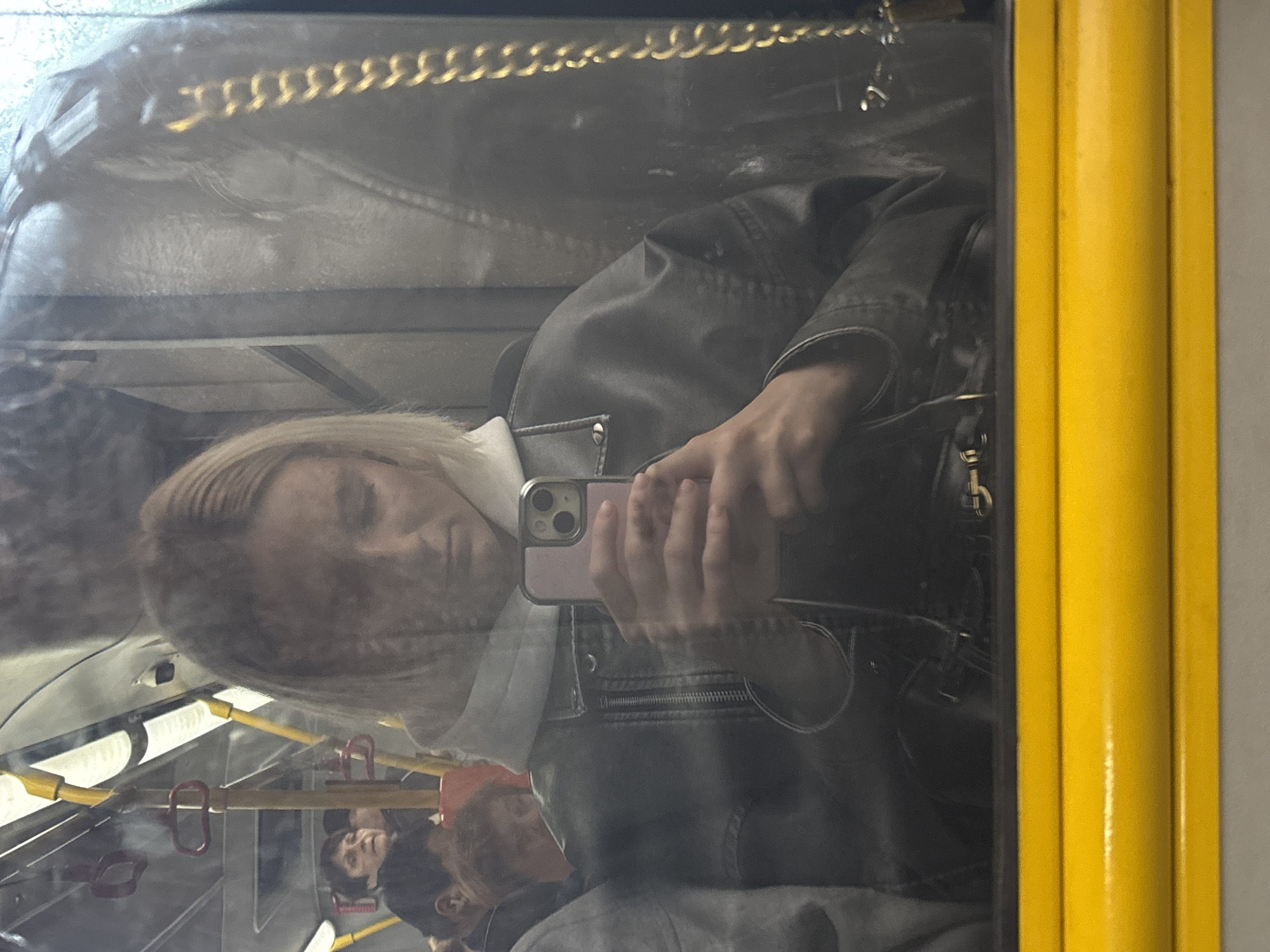 A photograph taken inside a public transportation vehicle in Saint Petersburg, Russia, captures a reflection of a woman in the window. The woman is holding a smartphone, seemingly taking a selfie. She is wearing a jacket and a light-colored shirt or turtleneck. In the reflection, other passengers can be seen, including at least two people seated behind her. The interior of the vehicle features yellow handrails and support structures. The image is framed by a yellow window frame. A bag with a chain strap is visible near the woman's hand.