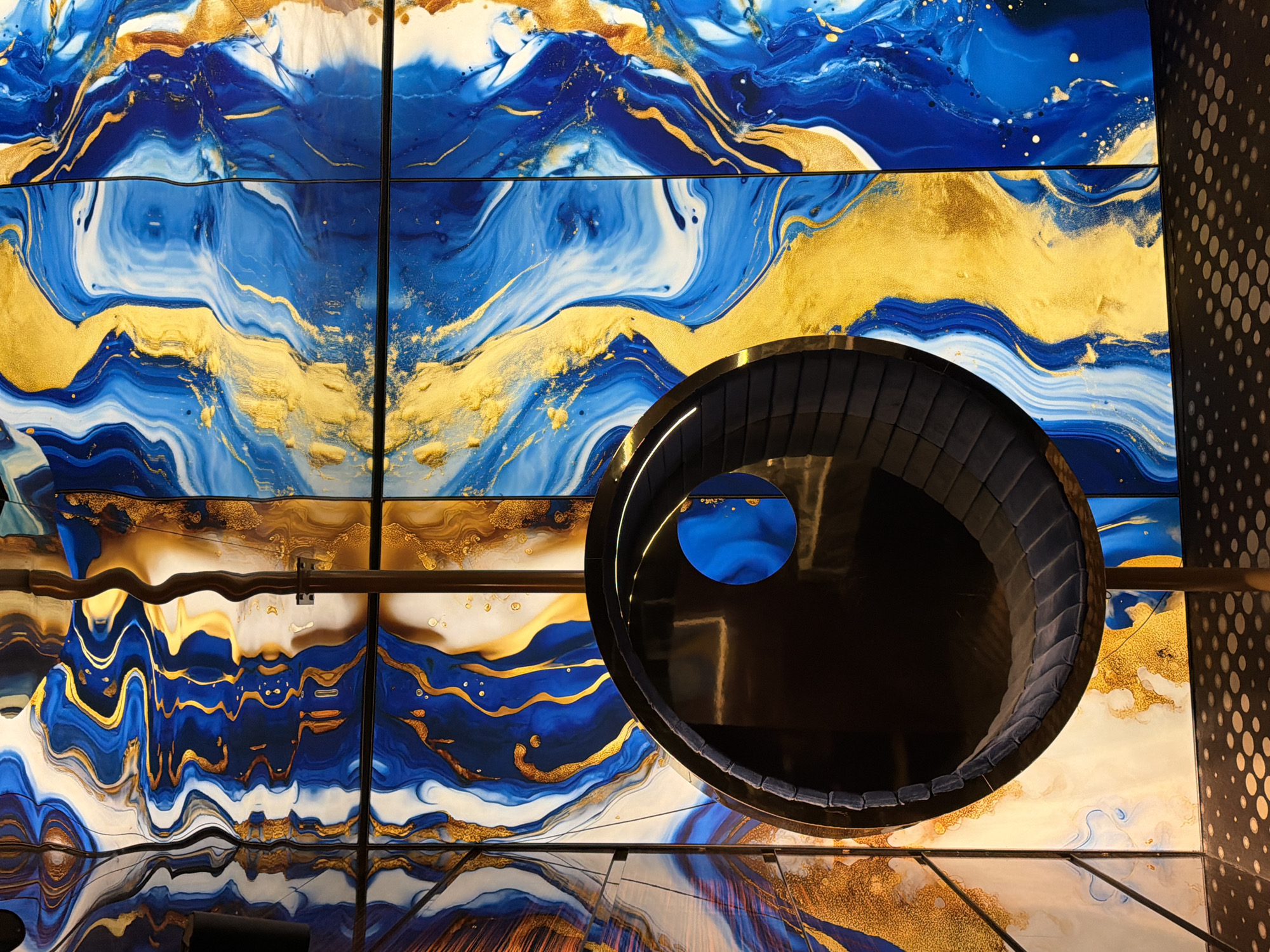 The image shows an interior architectural detail. A round, black, upholstered seating structure is positioned near a ceiling comprised of square panels displaying an abstract design of swirling blue, white, and gold patterns reminiscent of liquid marble. The panels are illuminated from behind. The lower portion of the image suggests a mirrored or glossy surface reflecting the ceiling design. A section of a patterned wall with dark circles is visible on the right side. The exact location in Varna, Bulgaria, cannot be determined from the image alone.
