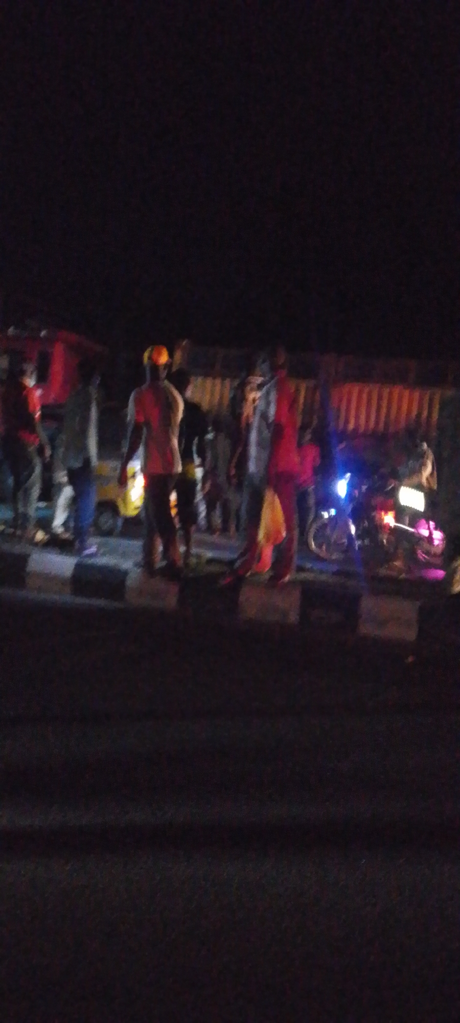 At night in Unknown, Unknown, there is a gathering of several individuals near a roadway. A vehicle is present, along with a motorcycle that has lights illuminated. The area appears to be near a street with a curb, and a barrier is visible in the background. Some of the individuals are wearing light-colored clothing. The lighting is poor.