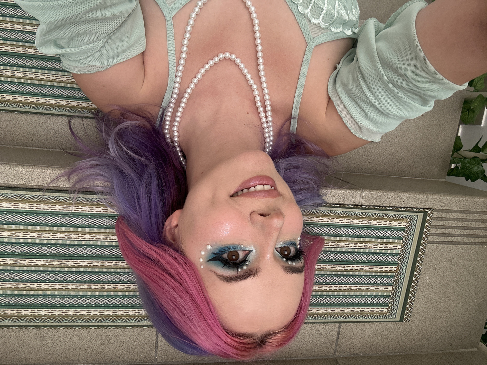 A woman with dyed purple and pink hair is lying down on a patterned rug, likely indoors. She is wearing a light green top and a pearl necklace. Her makeup includes blue eyeshadow and pearl accents around her eyes. The surrounding environment appears to be on steps, with a textured rug and decorative plants visible. It is not possible to determine the location within Zagreb, Croatia, from the image alone.