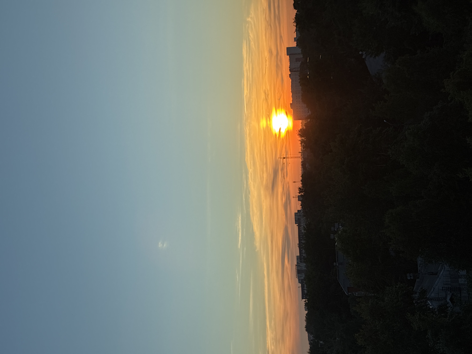 A sunset paints the sky over a city skyline. The sun, a bright orb of yellow and white, is positioned low in the sky, casting a warm glow across the horizon. Wispy clouds, tinged with orange and pink, stretch across the sky, creating a beautiful gradient. In the foreground, silhouetted against the vibrant sky, are the dark shapes of trees and buildings. The trees form a dense canopy, obscuring much of the lower cityscape. Among the trees, the tops of buildings are visible, their details softened by the fading light. A few faint lights can be seen in some of the structures. The sky transitions from a clear, light blue at the top of the frame to a warm, orange hue near the horizon. The overall scene captures a peaceful moment as day turns to night.